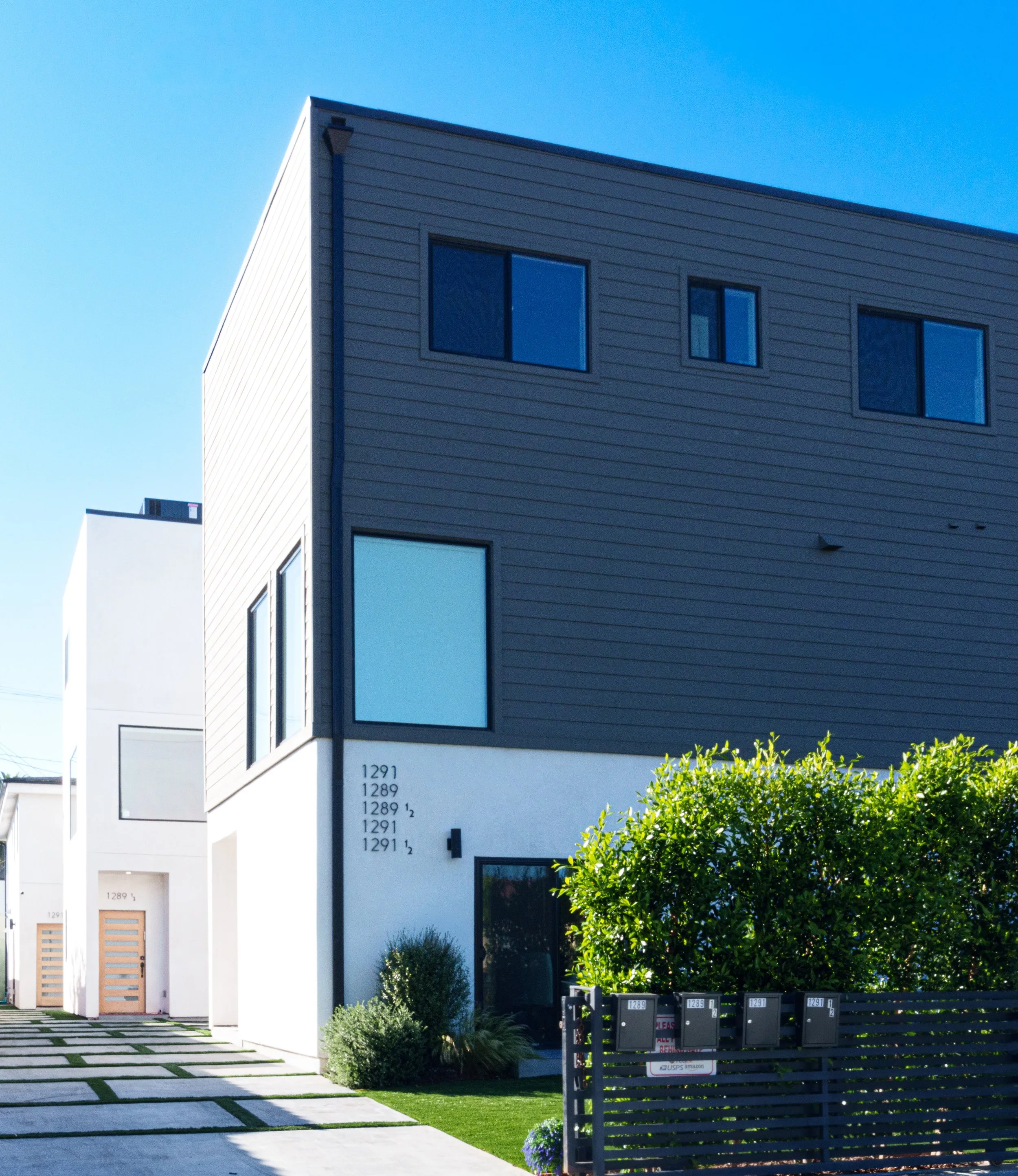 Modern multi-story residential building with white and dark gray exterior, large windows, and a small front yard with greenery and mailboxes.