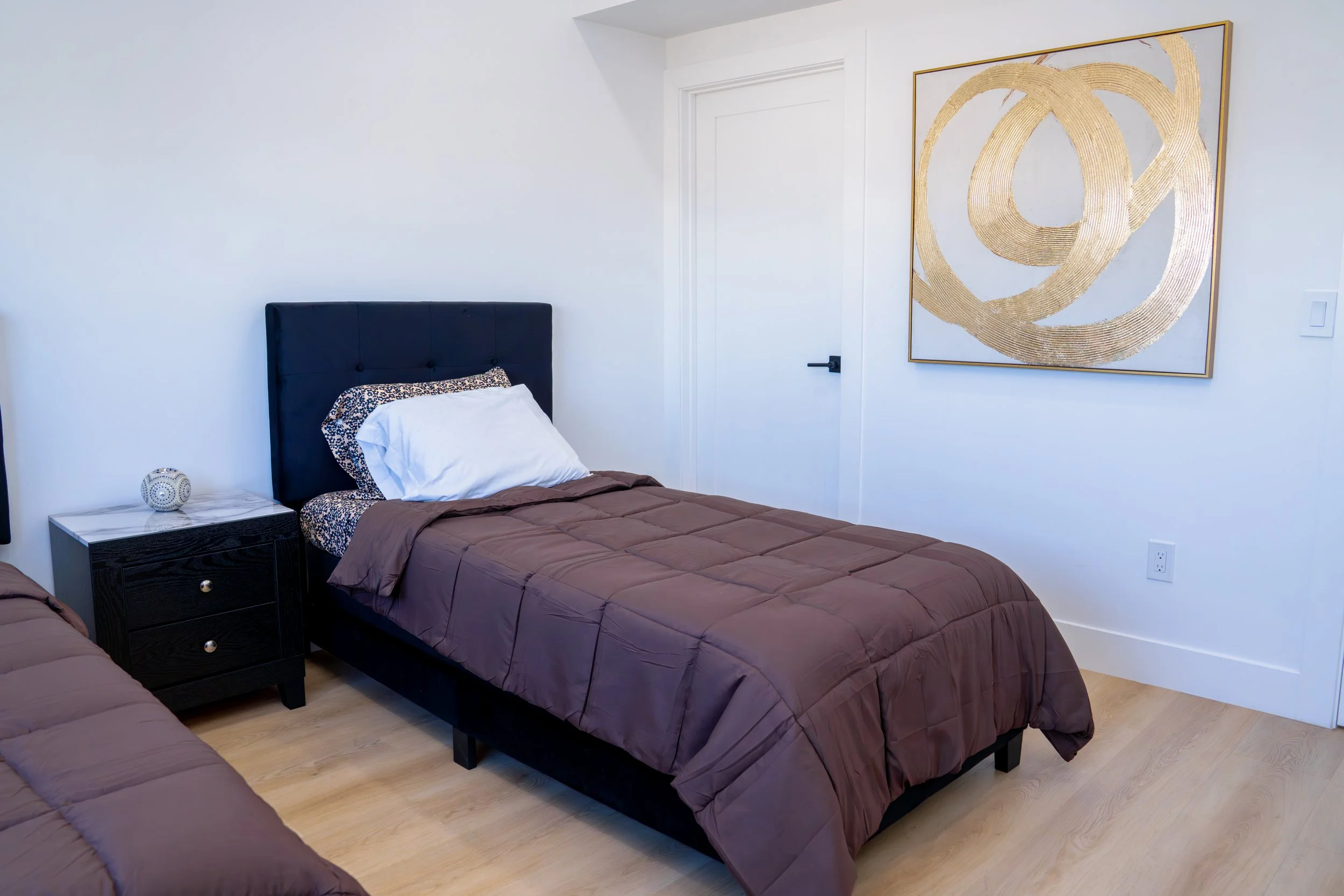 A bedroom with two beds covered in brown quilts. There is a black nightstand with three drawers between the beds, topped with a round decorative orb. A leopard-print pillow and a white pillow are on the bed in the foreground. The wall features a larg