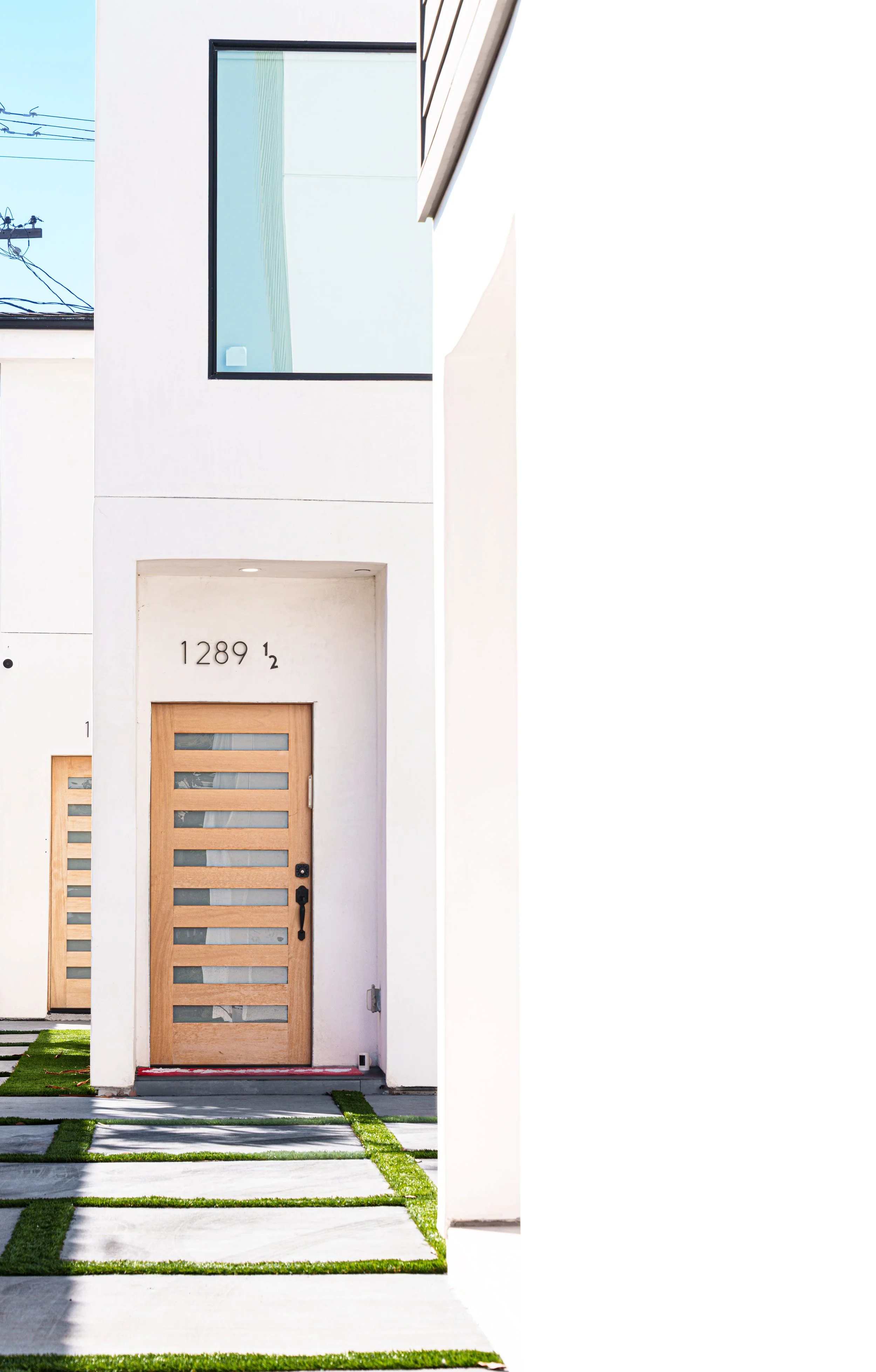 Modern white house with a wooden front door featuring horizontal glass panels, a number 1289 and a small number 1/2 above the doorway, and minimalist concrete walkway with grass strips.