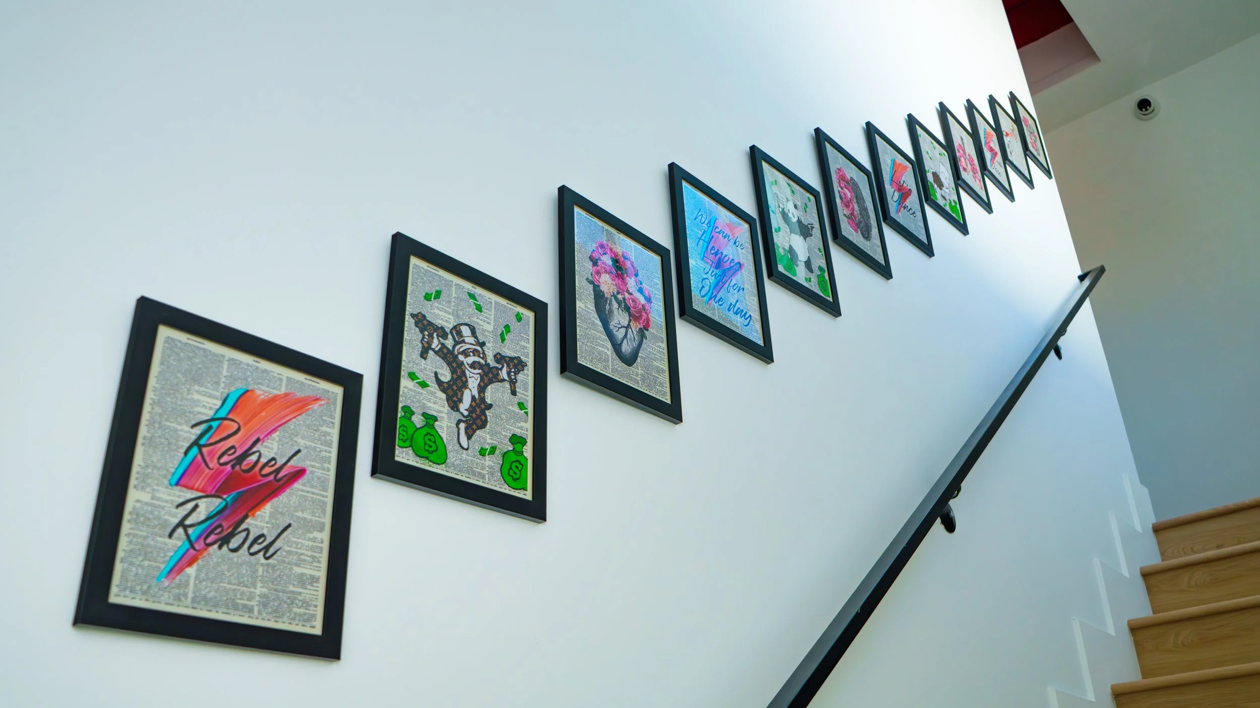 Gallery wall with framed artwork on a white wall, viewed from below and to the right, with a staircase visible on the right side.