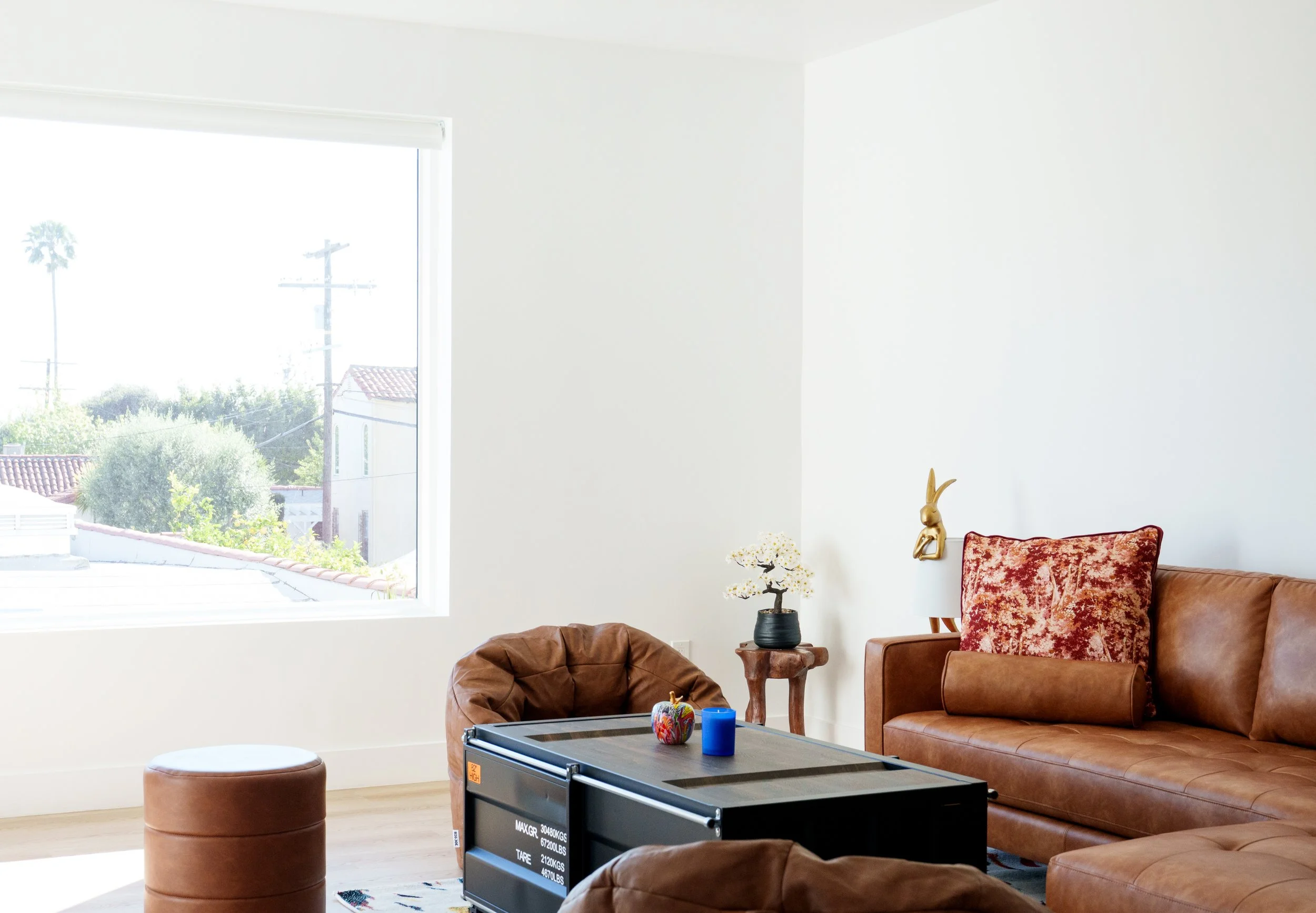 Living room with large window, brown leather sofa and matching ottoman, black coffee table with candles and decorative items, small side table with a bonsai tree, white wall-mounted lamp with bunny-shaped gold decoration, and bright natural light.