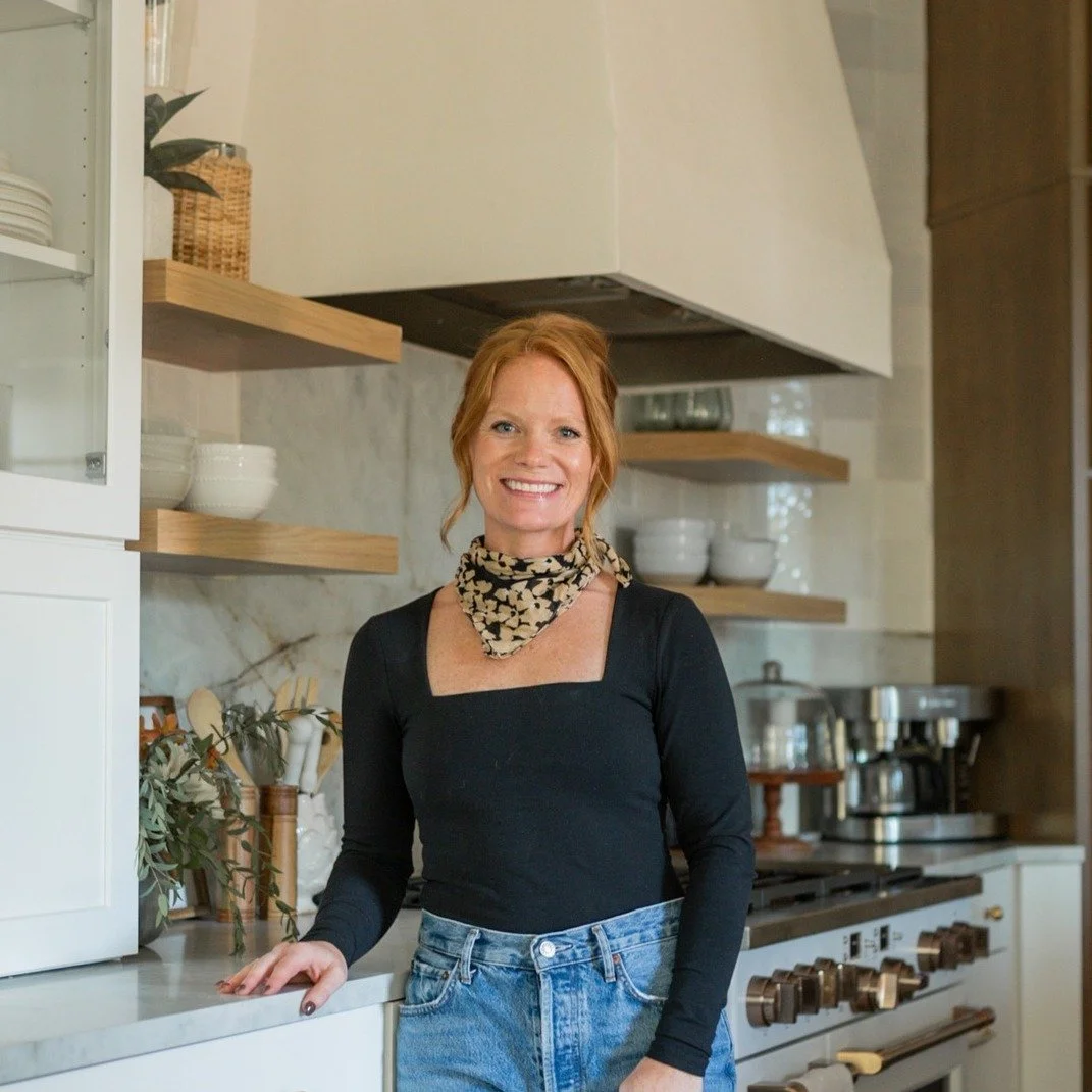A woman with red hair, wearing a black top with a square neckline, a patterned scarf, and blue jeans, stands in a modern kitchen smiling.