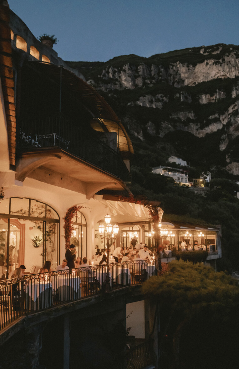 Outdoor restaurant terrace at night with tables, chairs, and people dining, decorated with string lights and greenery, overlooking a mountain landscape.