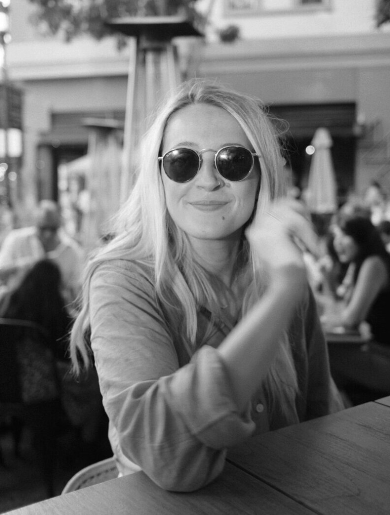 A woman with long blonde hair wearing round sunglasses sitting at a table outdoors in a busy patio area, smiling at the camera.