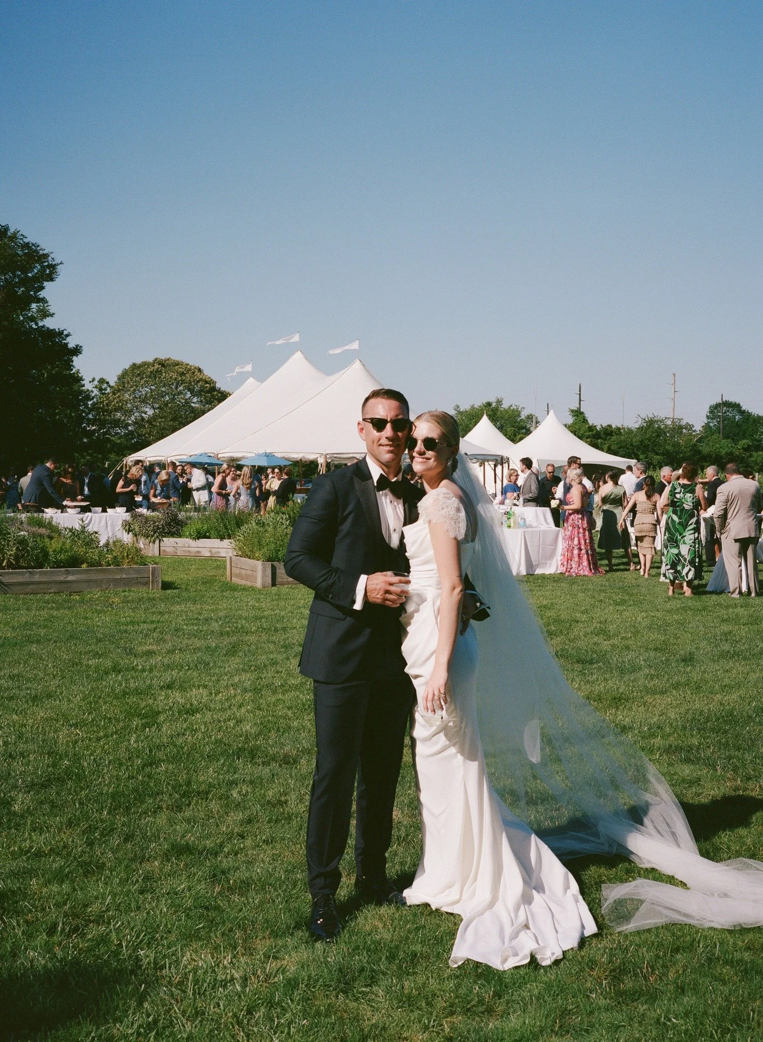 A couple dressed in wedding attire standing on a green lawn at an outdoor wedding reception with white tents and guests in the background on a sunny day.