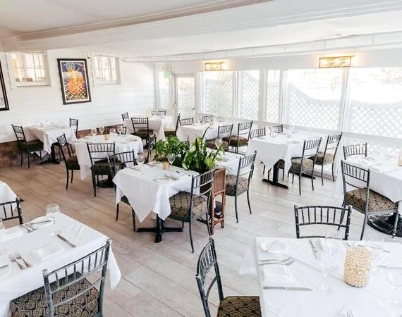 Restaurant dining room with white tablecloths, chairs, and place settings, decorated with plants and art on the walls.