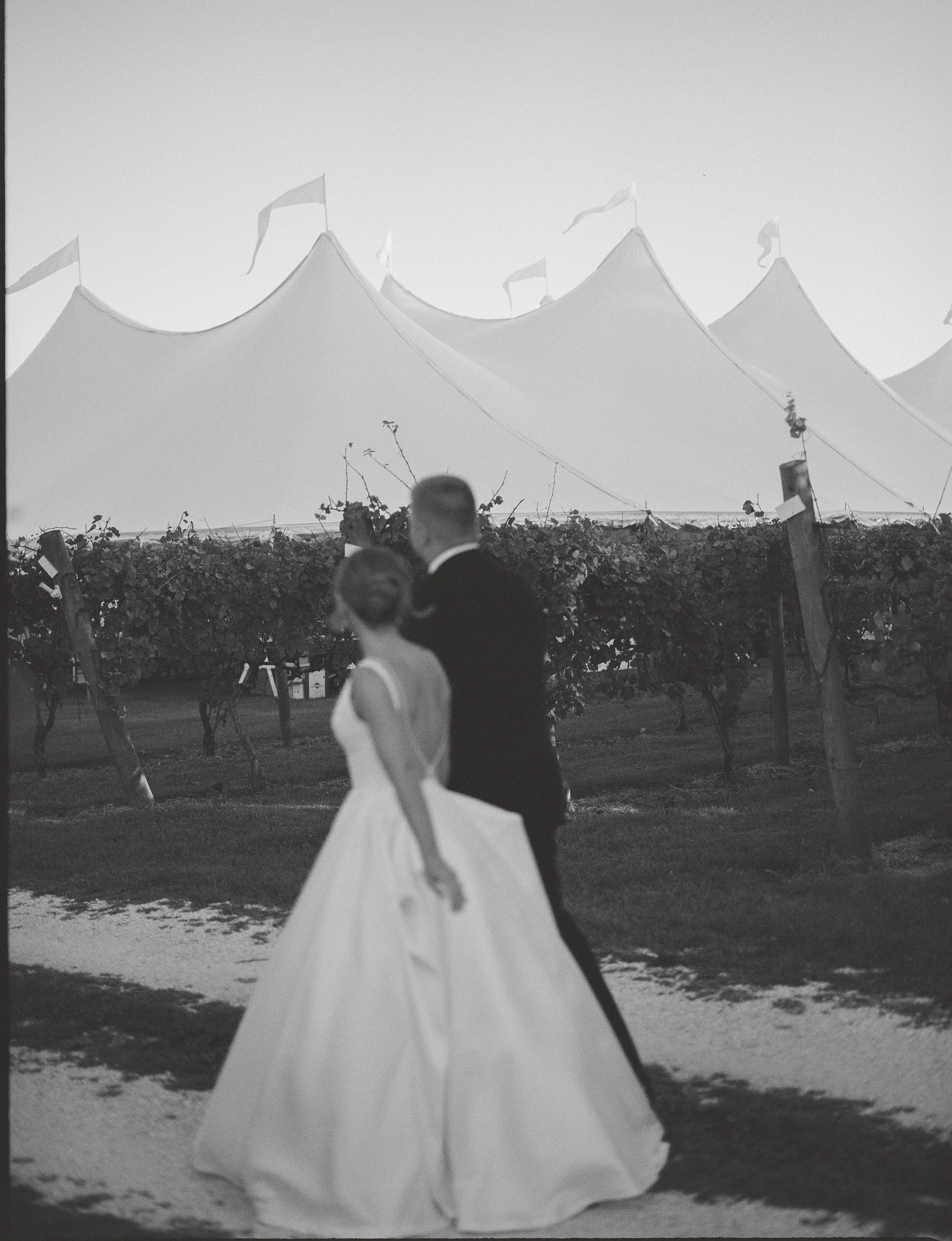 A couple dressed in formal wedding attire, standing outdoors under a large tent with flags, holding hands and walking in a vineyard or garden.