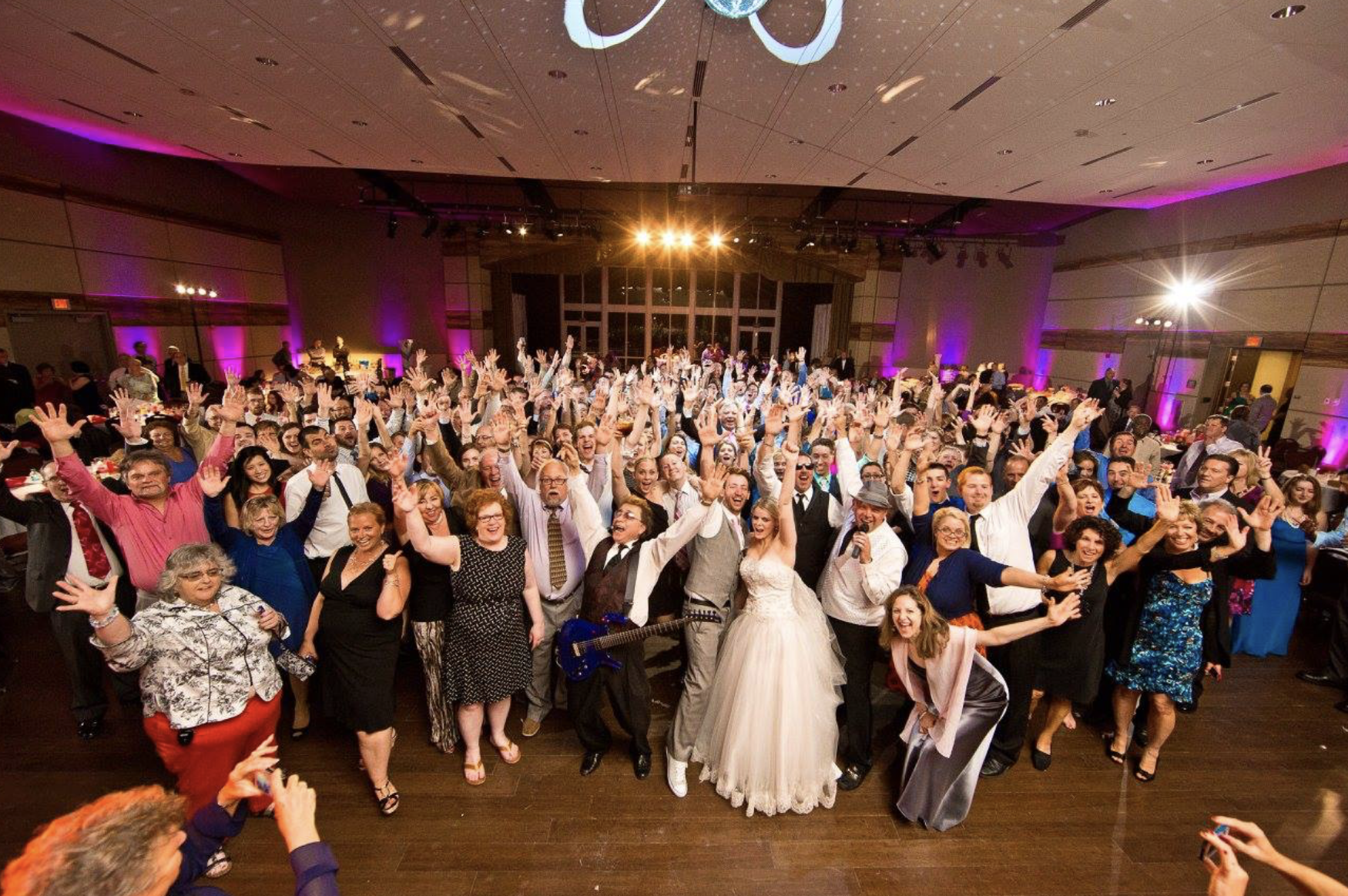 Large group of people dancing and celebrating at a wedding reception with purple and pink lighting.
