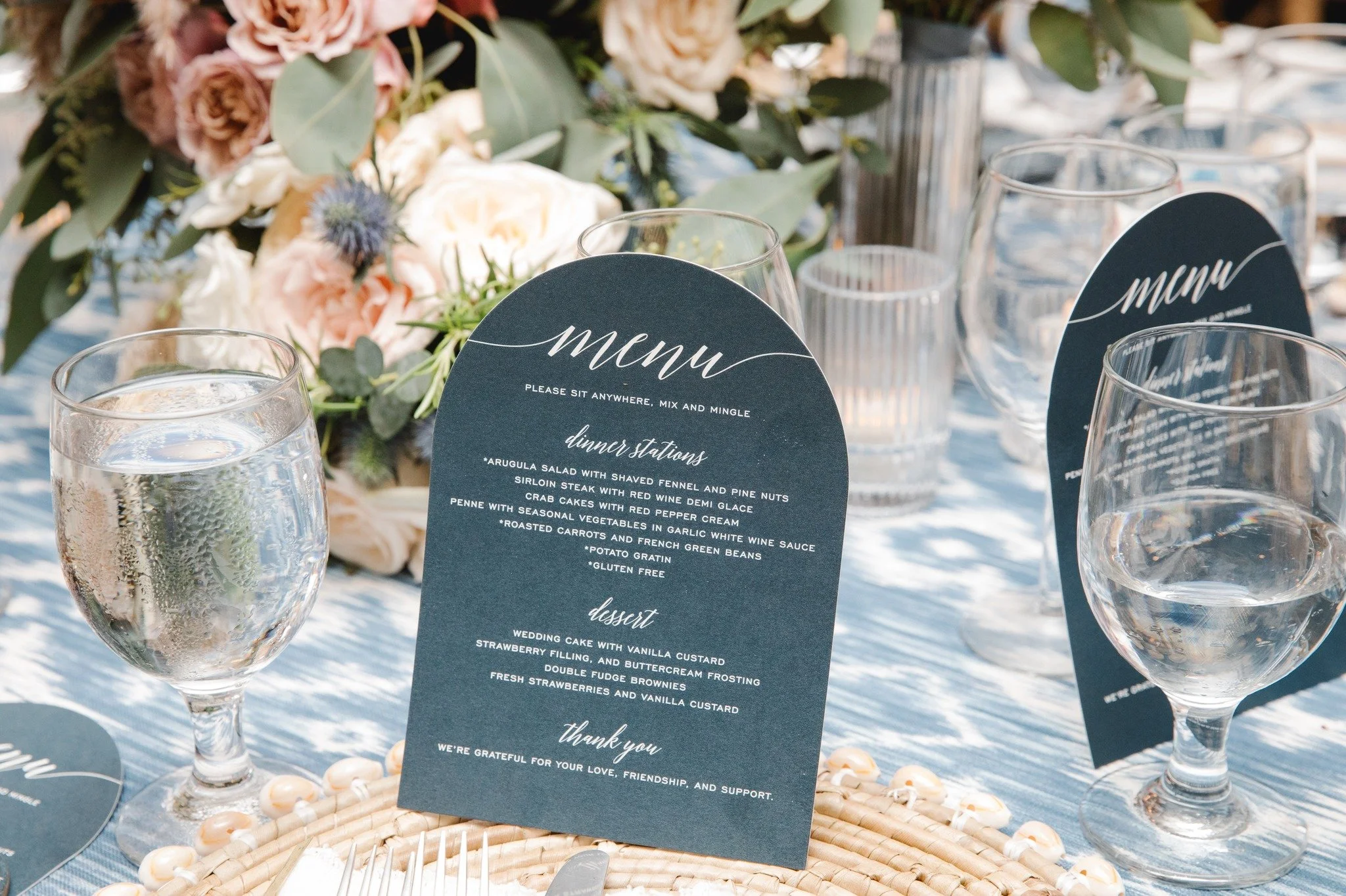 A wedding reception table setting featuring a printed menu card, surrounded by wine glasses and floral arrangements with roses and greenery.