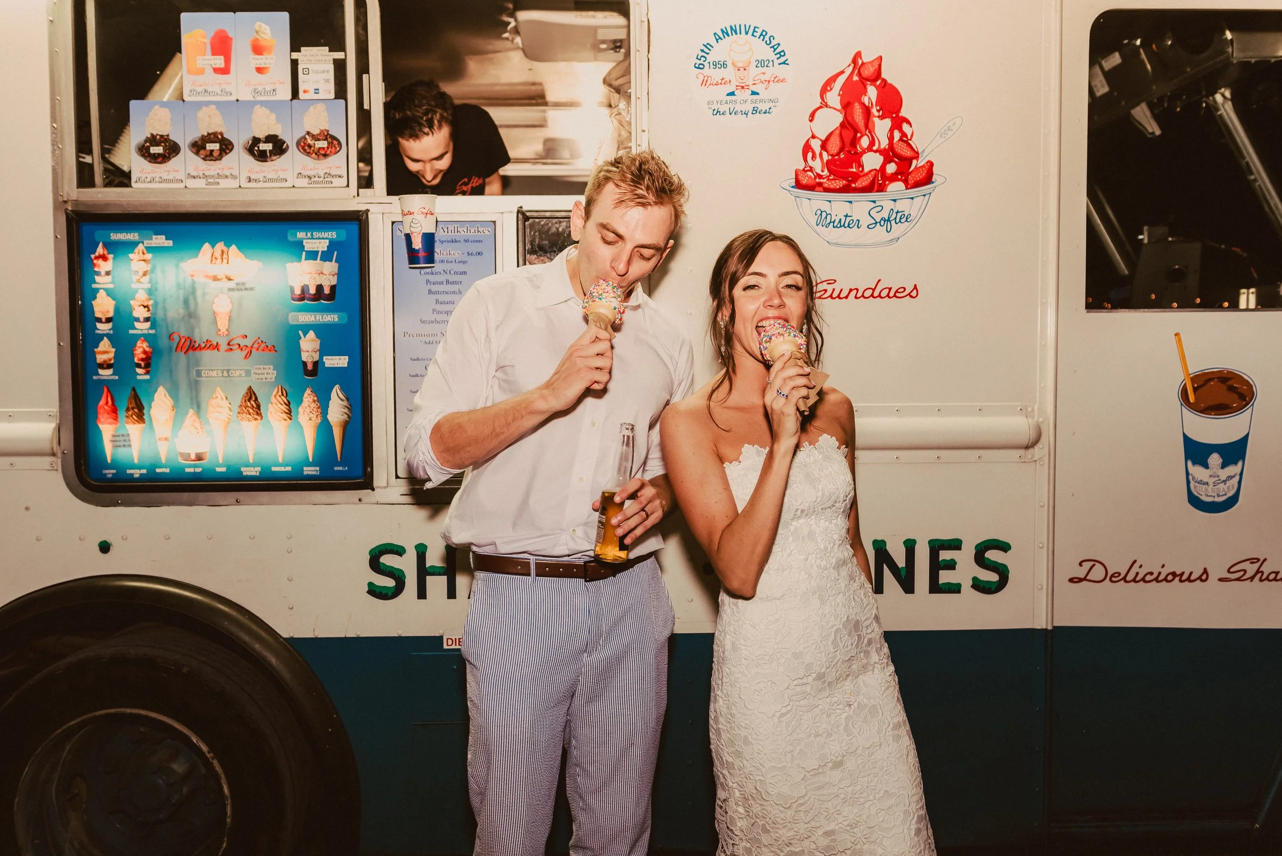 A man in a white shirt and striped pants and a woman in a white lace dress sharing ice cream cones in front of an ice cream truck, with a happy expression.