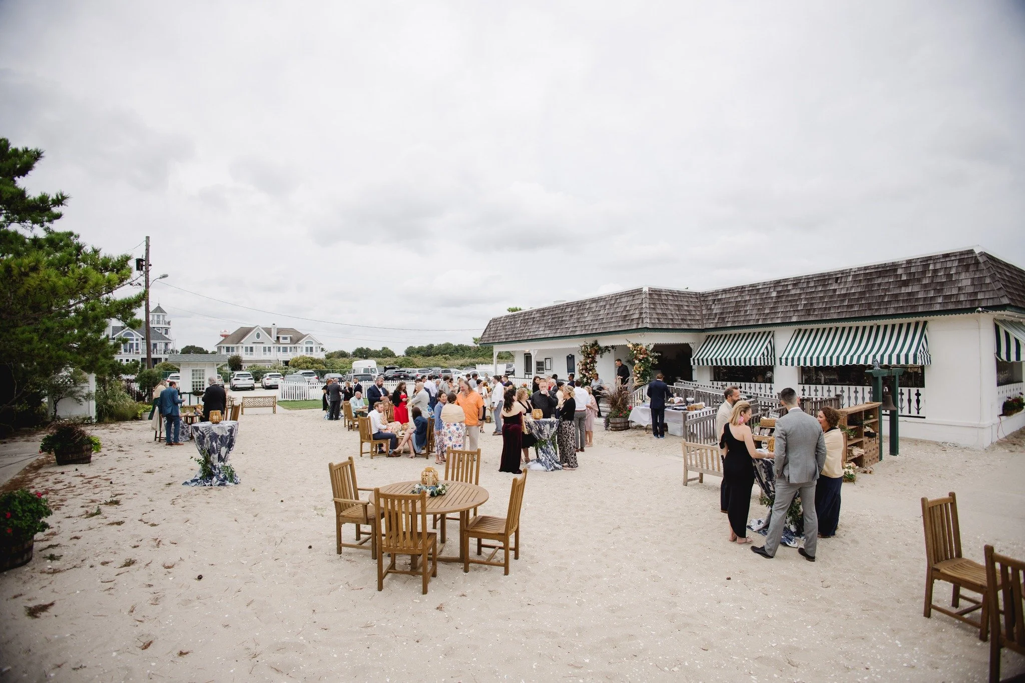 Outdoor wedding reception gathering with guests mingling and tables set up on a sandy surface, adjacent to a white building with striped awnings, under a cloudy sky.