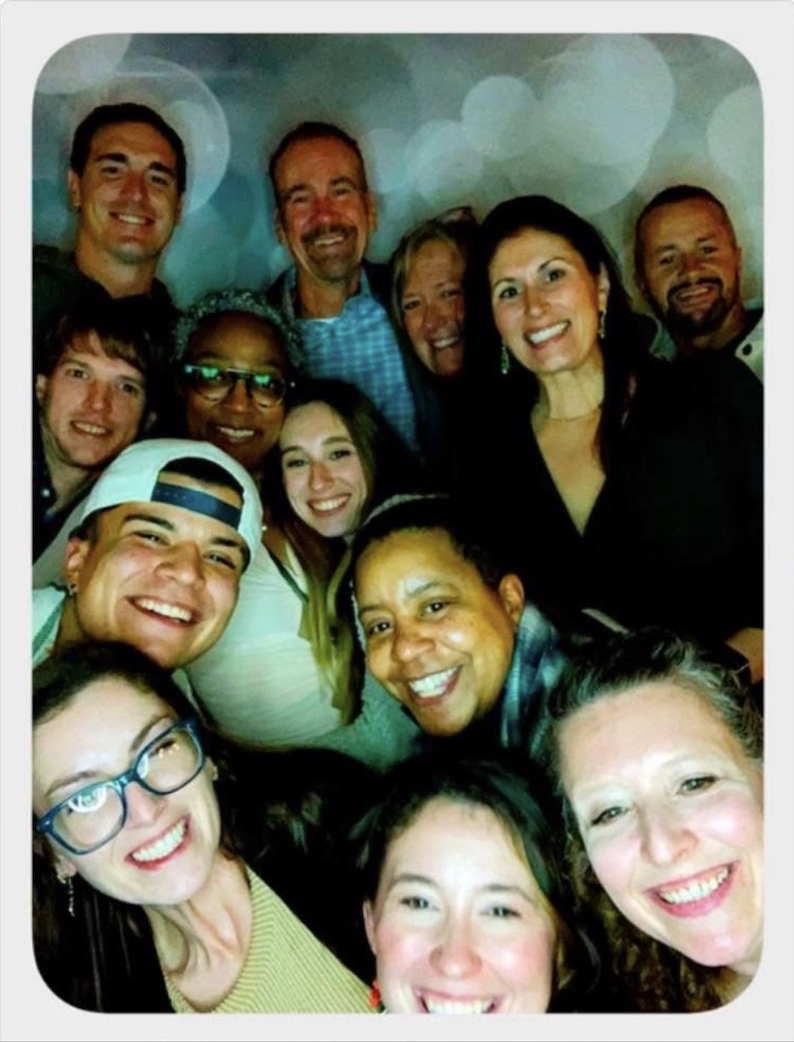 Smiling group of people taking a selfie together at a social gathering, with colorful bokeh lights in the background.