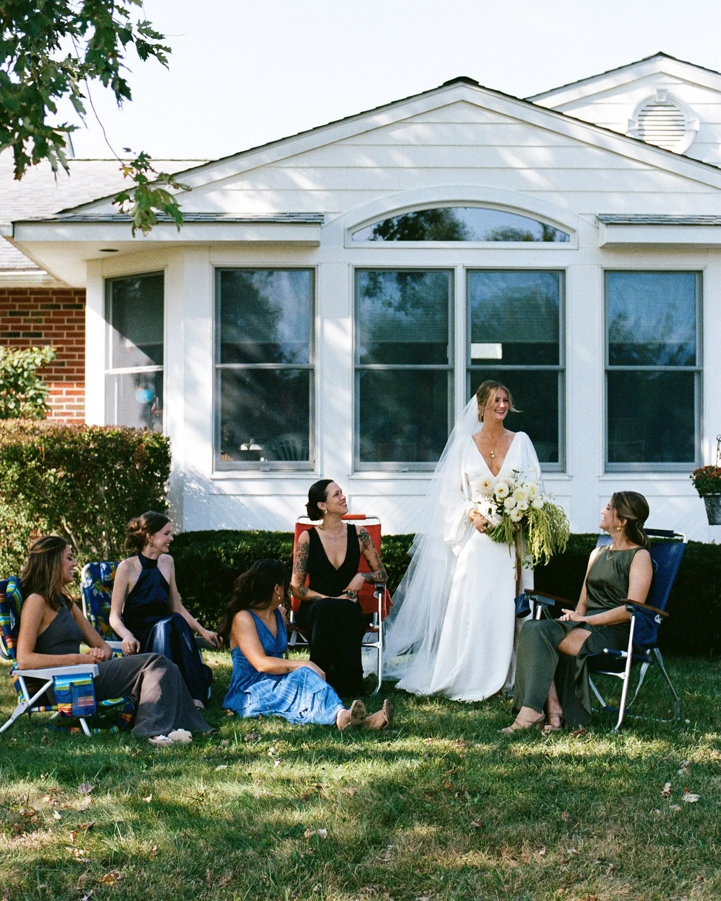 A group of women outside in a garden, with a woman in a white wedding dress holding a bouquet, smiling, surrounded by seated women in dresses and a woman in a dark blazer. Behind them is a house with a large window.