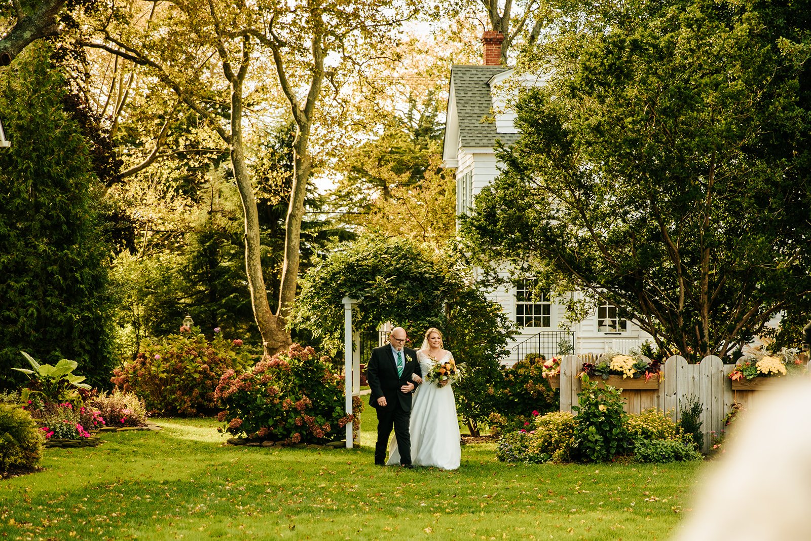 A bride and an older man, possibly her father, walking arm-in-arm in a garden with lush greenery and colorful flowers, next to a white house with a fenced flower bed, on a sunny day.
