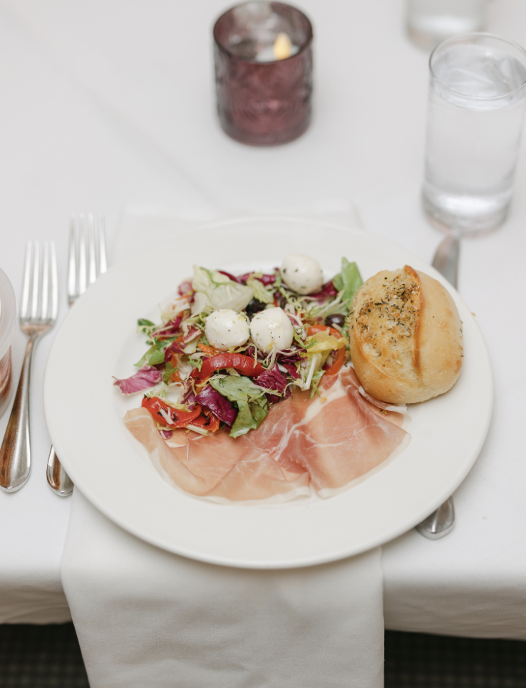 A plate of mixed greens salad with mozzarella balls, slices of prosciutto, and a bread roll, served with a glass of water at a dining table.