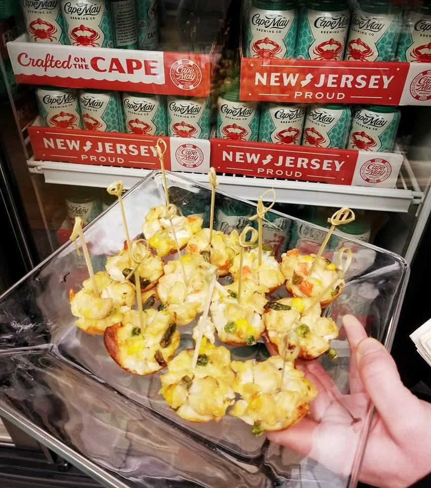 Finger foods, possibly stuffed or baked, topped with vegetables and cheese, skewered with small sticks, in a glass dish, in front of a cooler with Cape May Brewing Company cans and New Jersey patriotic signage.