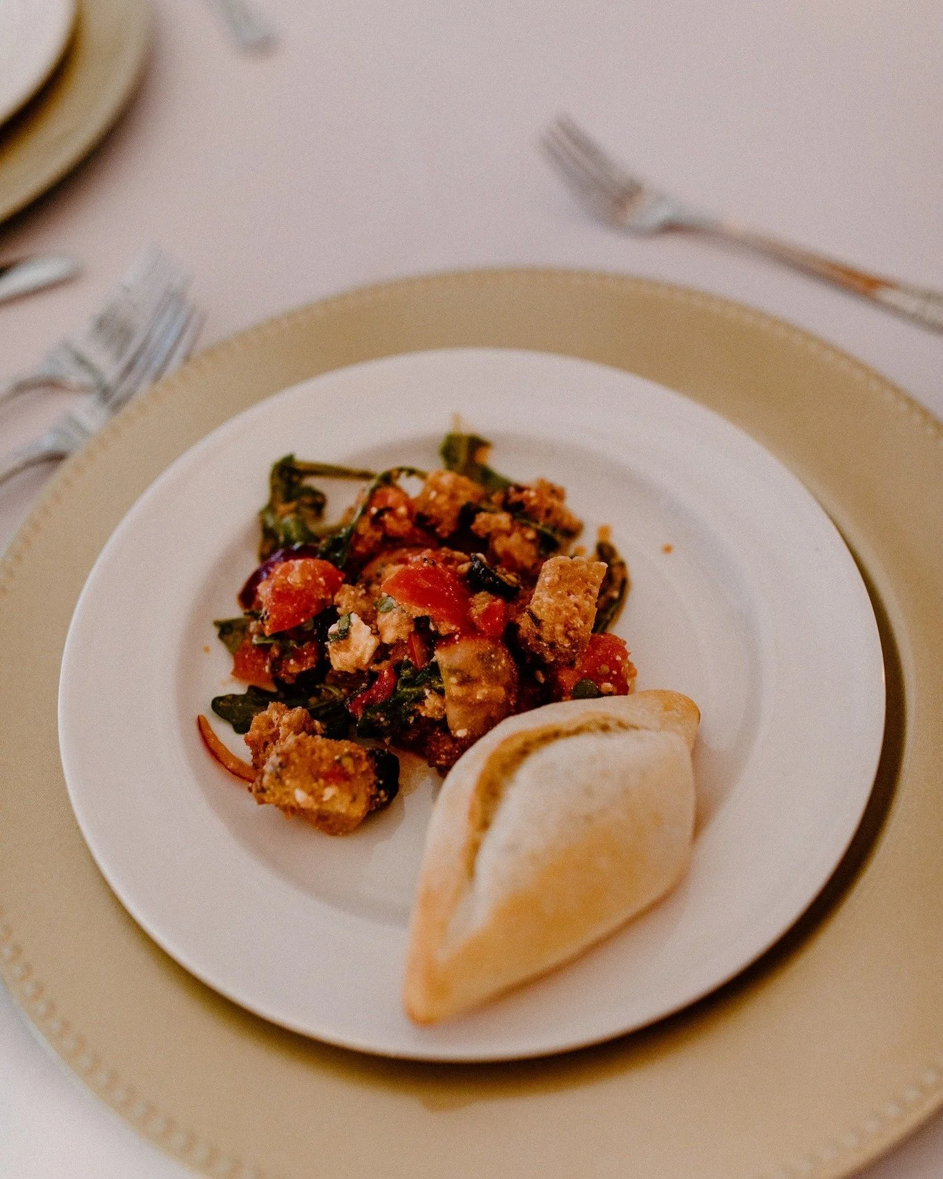 Close-up of a white plate with a mixed vegetable and protein dish, pieces of bread and two bread rolls, on a beige and gold placemat with silverware in the background.