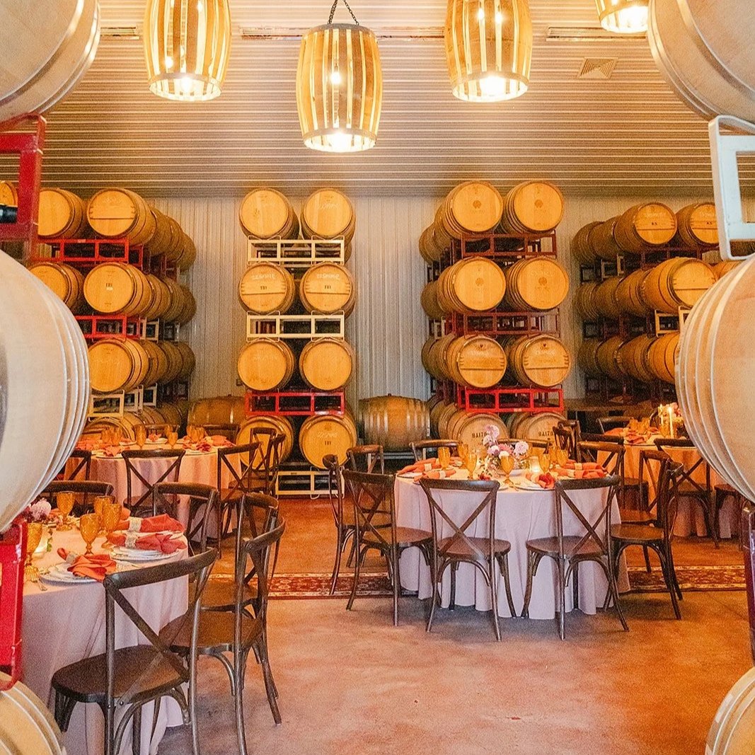 Wedding reception area with round tables, pink tablecloths, and chairs, set in front of stacked wine barrels in a rustic setting with warm lighting.