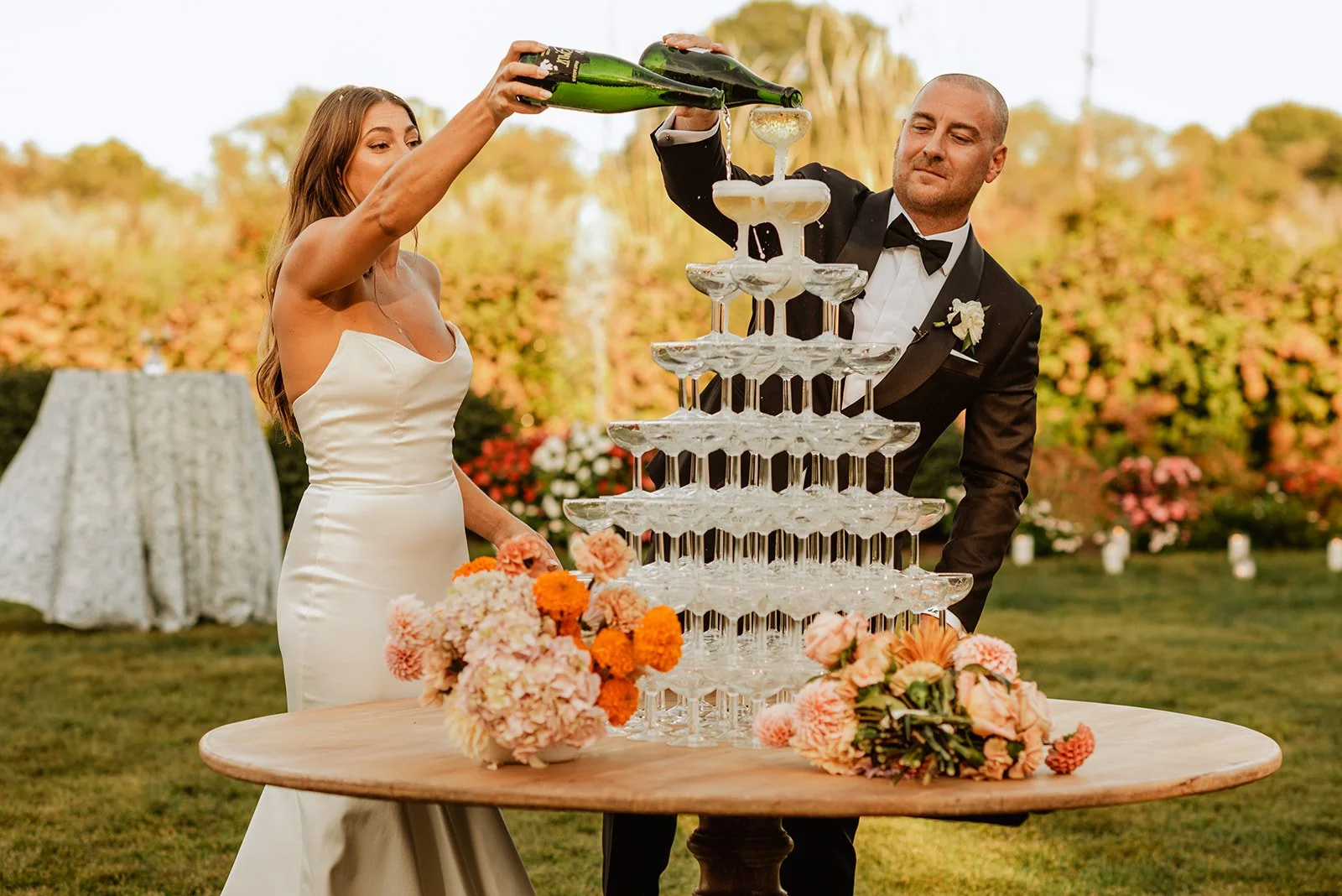 A bride and groom in wedding attire celebrate as they pour champagne into a multi-tiered champagne tower outdoors surrounded by flowers and greenery.