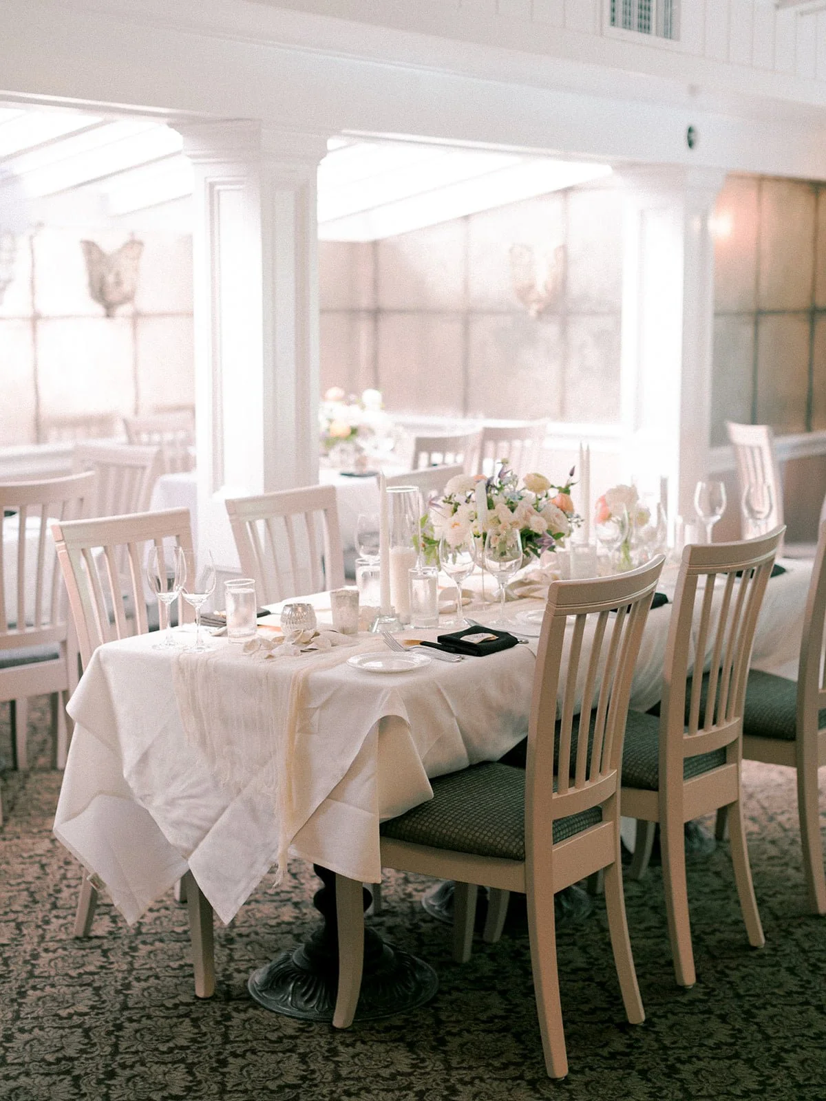 Elegant dining setup with a table covered in a white tablecloth, decorated with a floral centerpiece, candles, glassware, and silverware, in a well-lit room with white chairs around it.