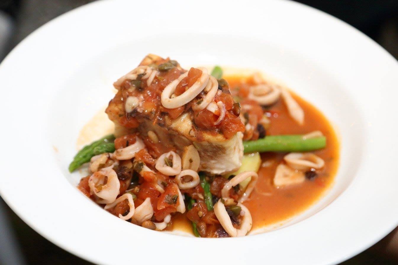 Plate of fish topped with tomato-based sauce and surrounded by vegetables and pasta.