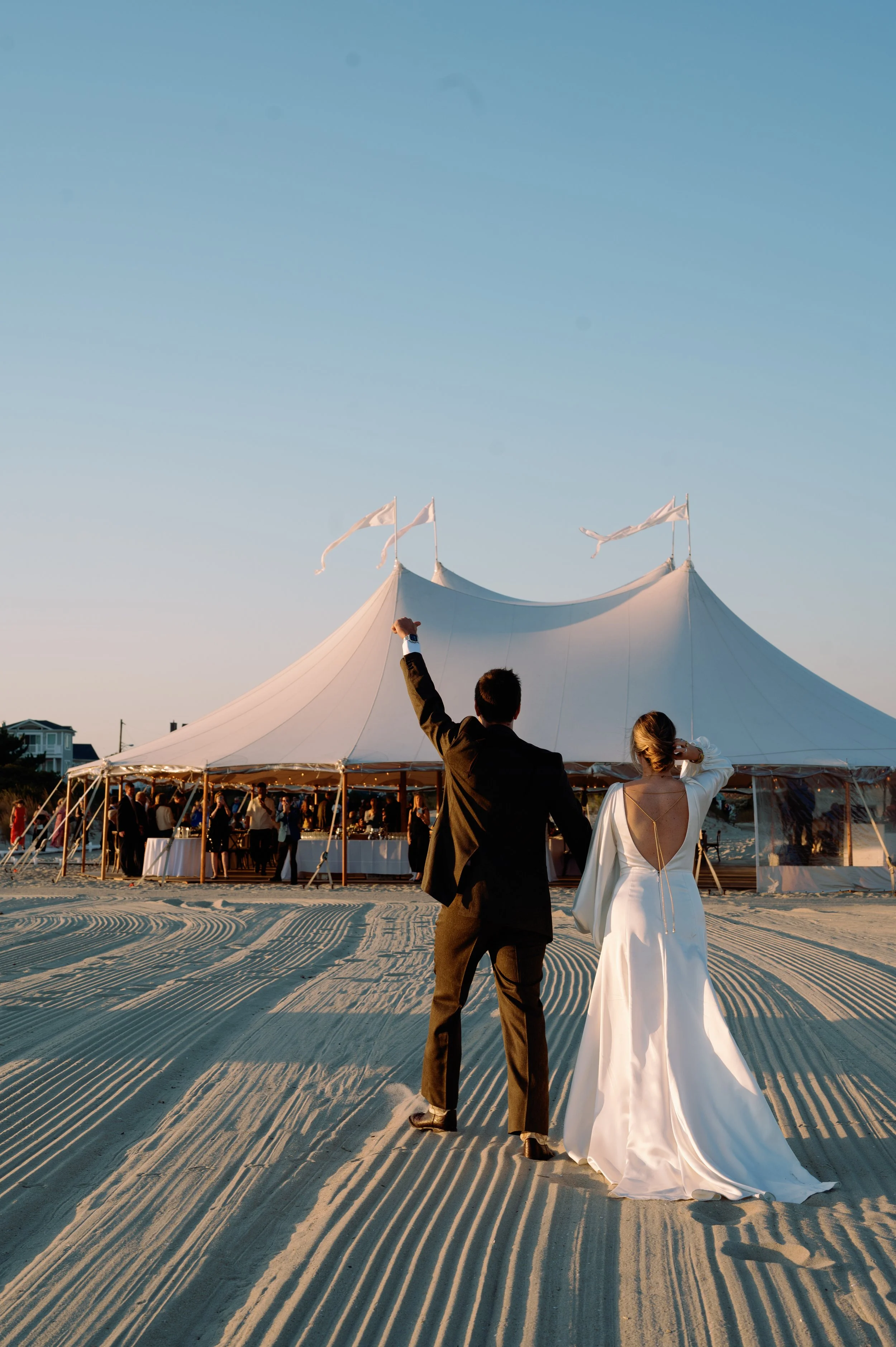 A bride and groom walk towards a large white wedding tent, holding their arms up in celebration, at sunset on a sandy beach with patterned sand dunes in the foreground and a clear blue sky.