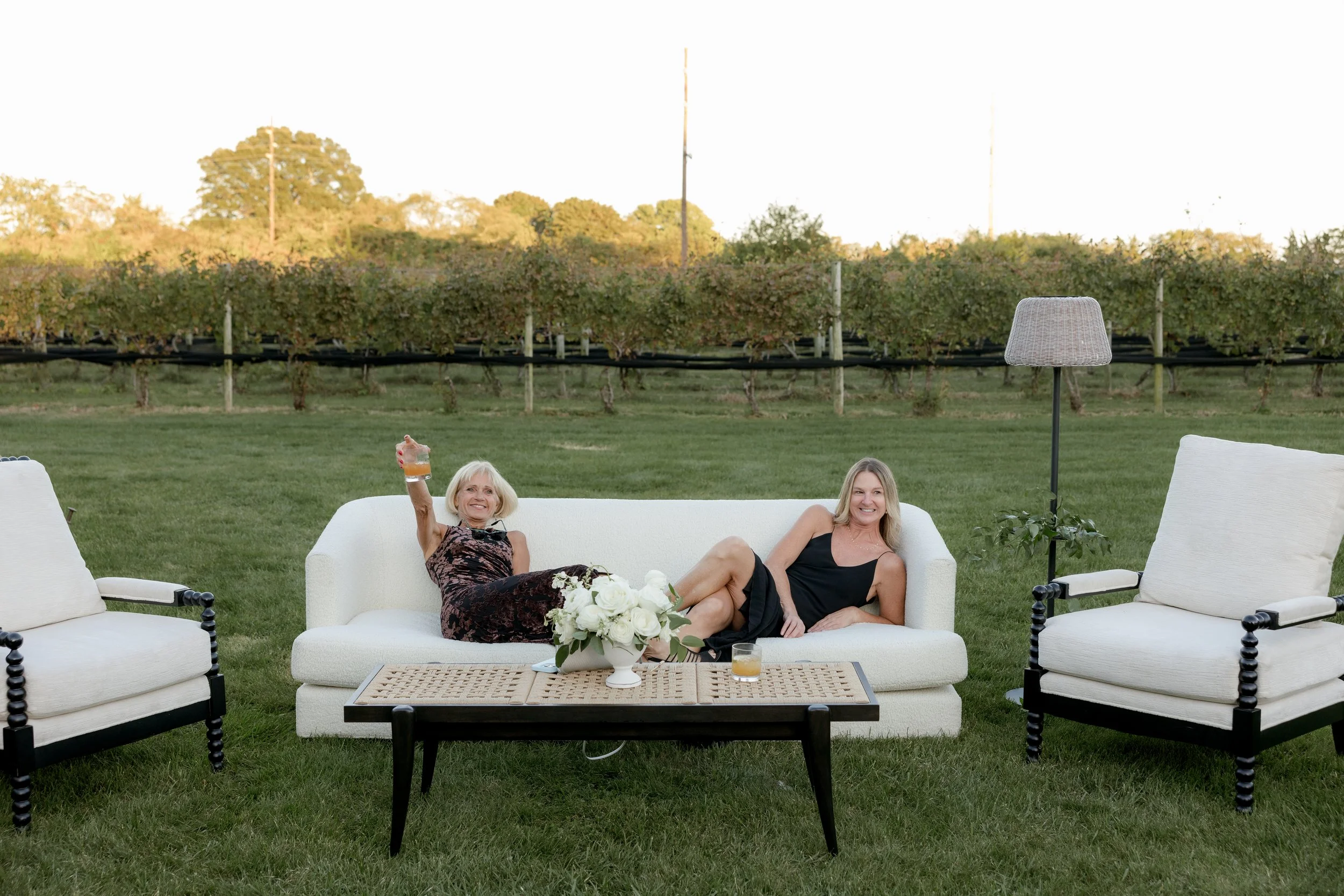Two women sitting on a white outdoor sofa in a grassy backyard, smiling and relaxing, with a table of white flowers and drinks in front of them, surrounded by white outdoor furniture and a vineyard in the background during sunset.