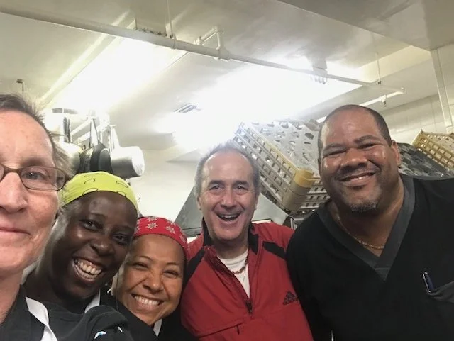 Group of five smiling people taking a selfie in a kitchen or food prep area.