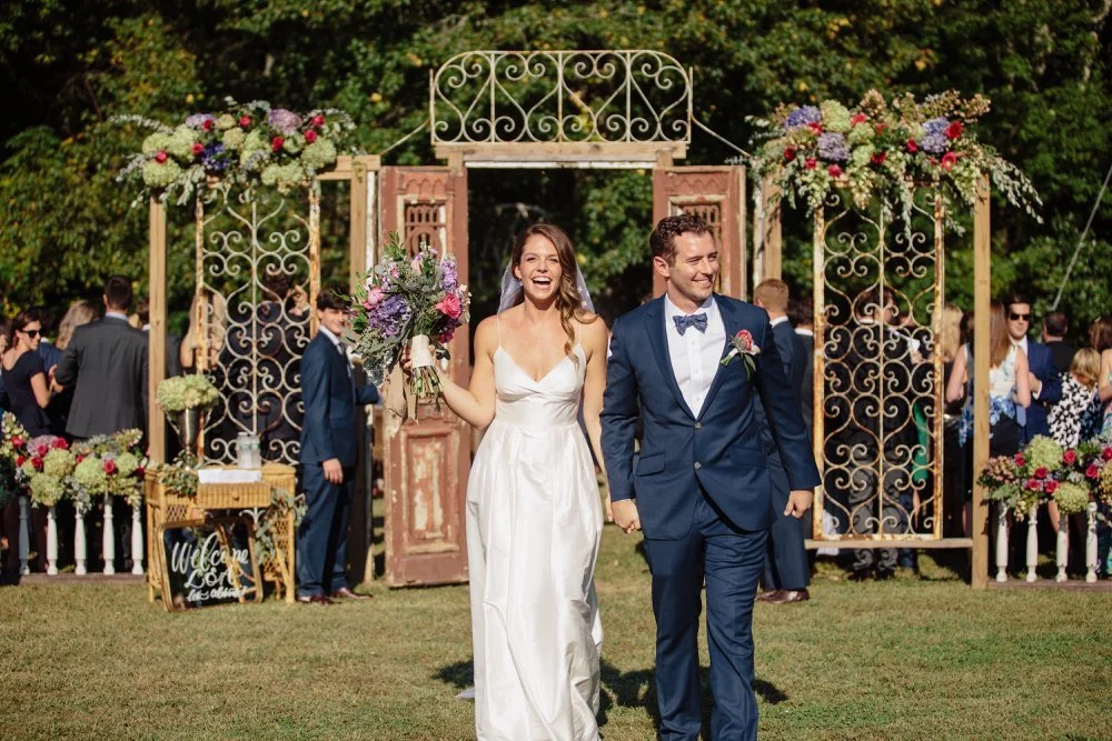 Bride and groom walking hand in hand at an outdoor wedding ceremony, surrounded by guests and floral decorations.
