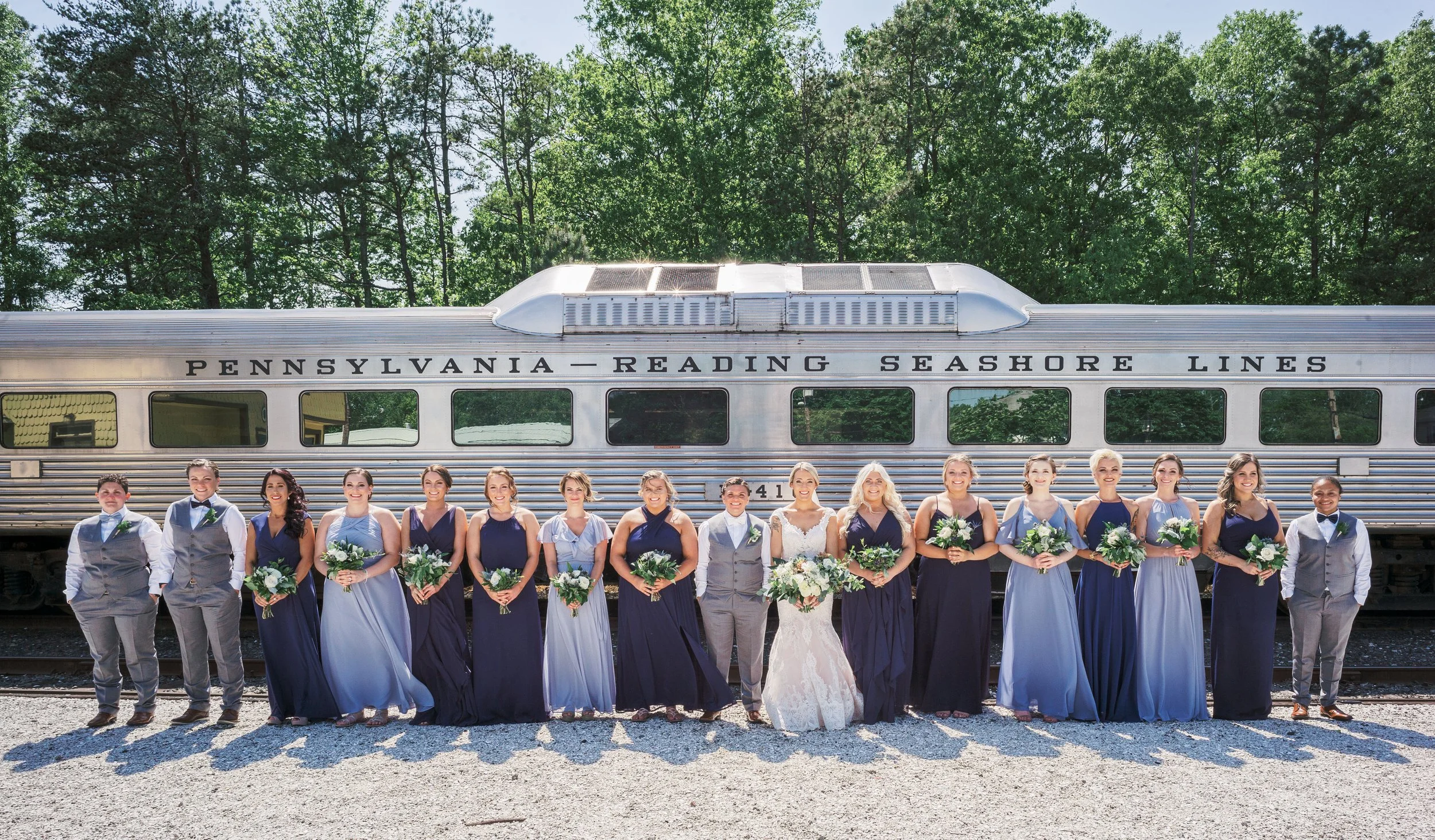 A group of bridesmaids and groomsmen standing in front of a silver train car labeled Pennsylvania Reading Seashore Lines, outdoors under trees, dressed in formal attire, holding bouquets, with a bride and groom in the center.