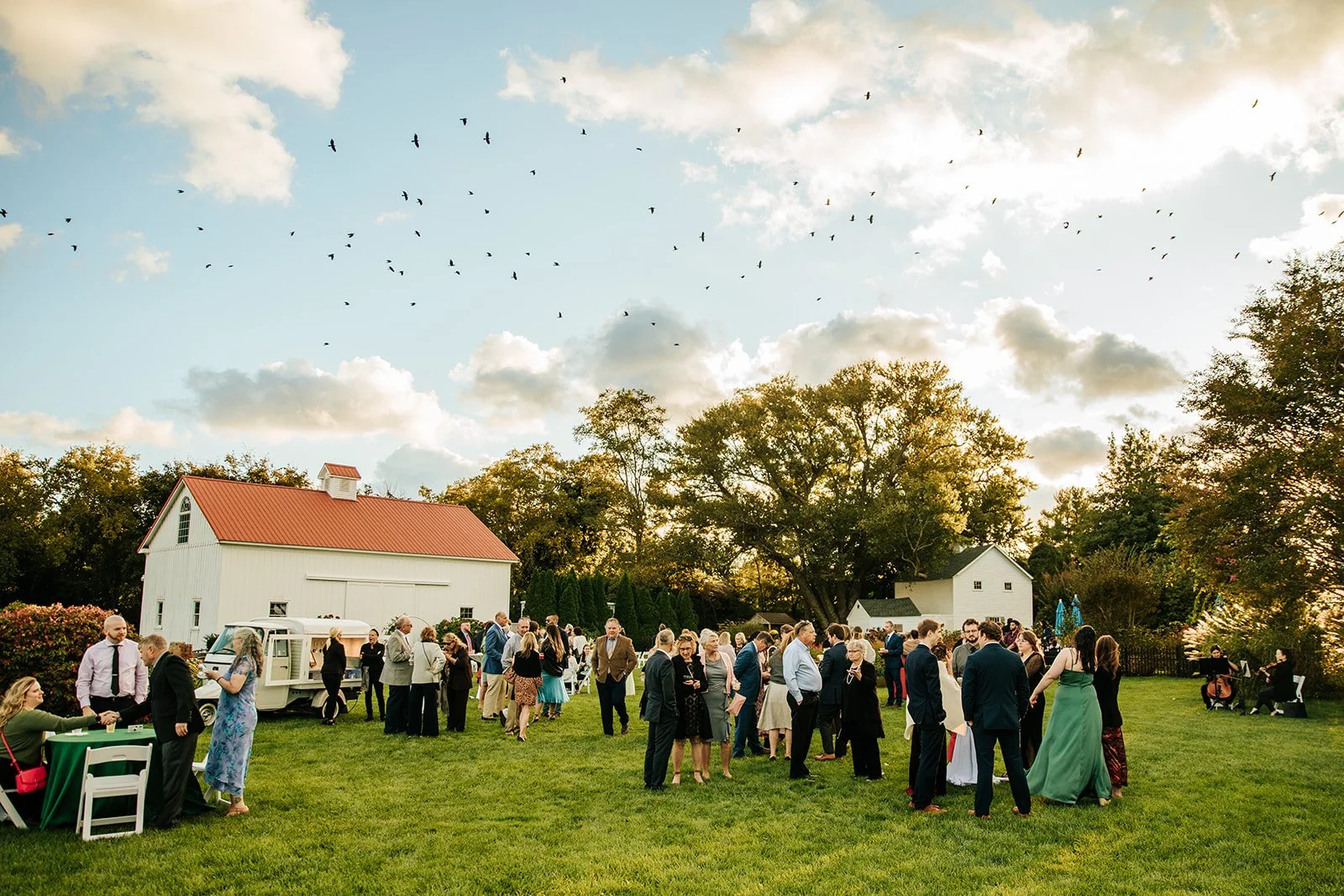 Group of people gathered outdoors on a lush green lawn during a social event, with a white barn and trees in the background under a partly cloudy sky during sunset.