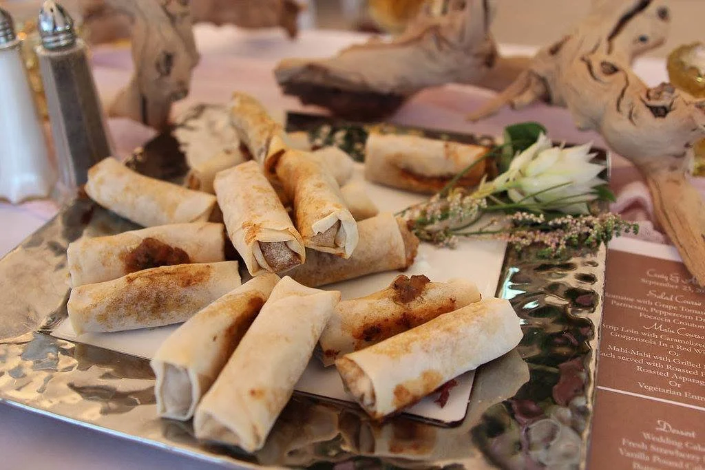 A silver tray with several rolled-up spring rolls, some with visible meat fillings. The tray also holds a small arrangement of white and purple flowers and green leaves, with a blurred menu in the background.
