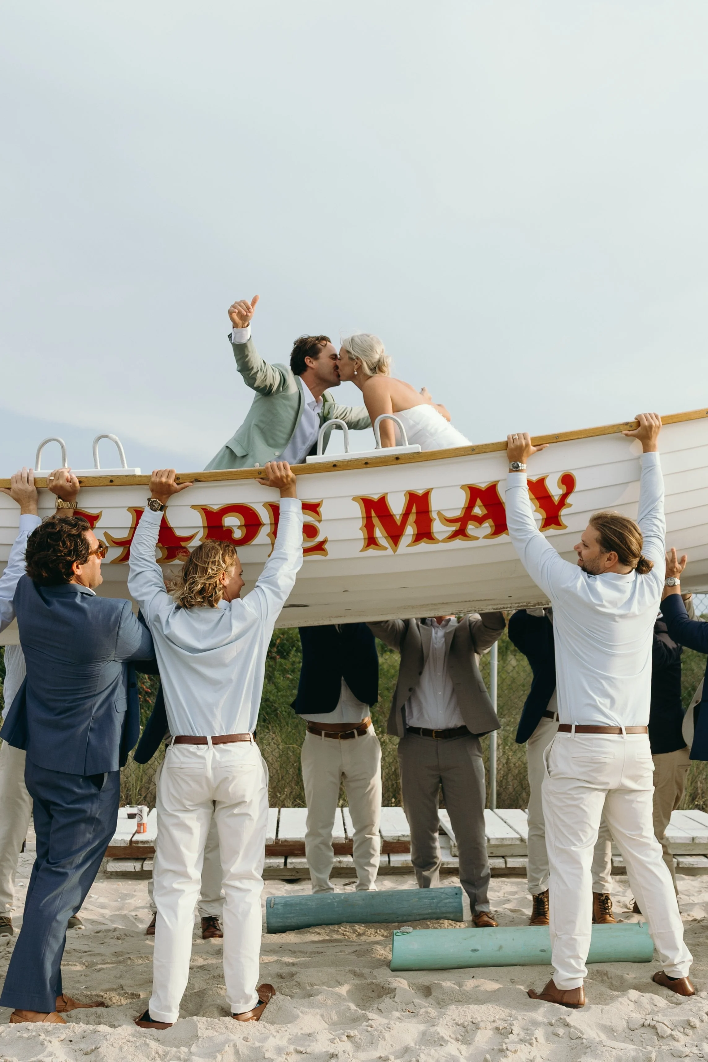 A newlywed couple kissing on top of a boat while friends lift the boat at a beach. The friends are dressed in formal suits, and the couple is dressed in wedding attire, with the bride in a strapless white gown and the groom in a light-colored suit. The boat has words painted on it, partially visible, and the sky is clear.