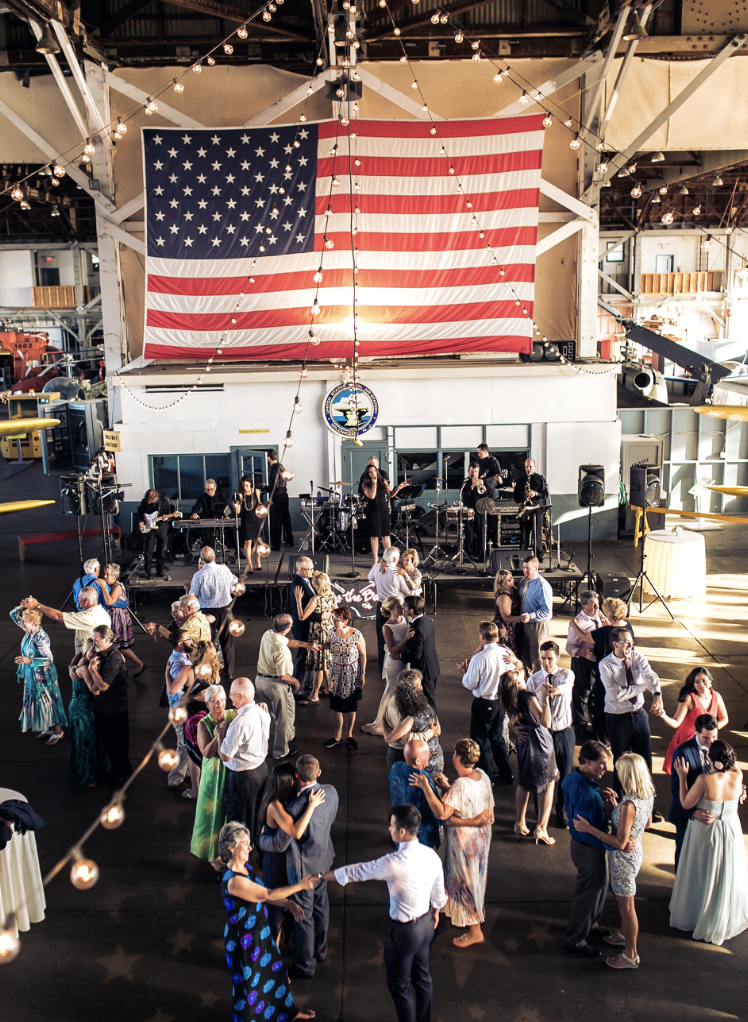 People dancing in a large indoor space decorated with string lights and a large American flag hanging on the back wall. A band is performing on a stage behind the dancers.