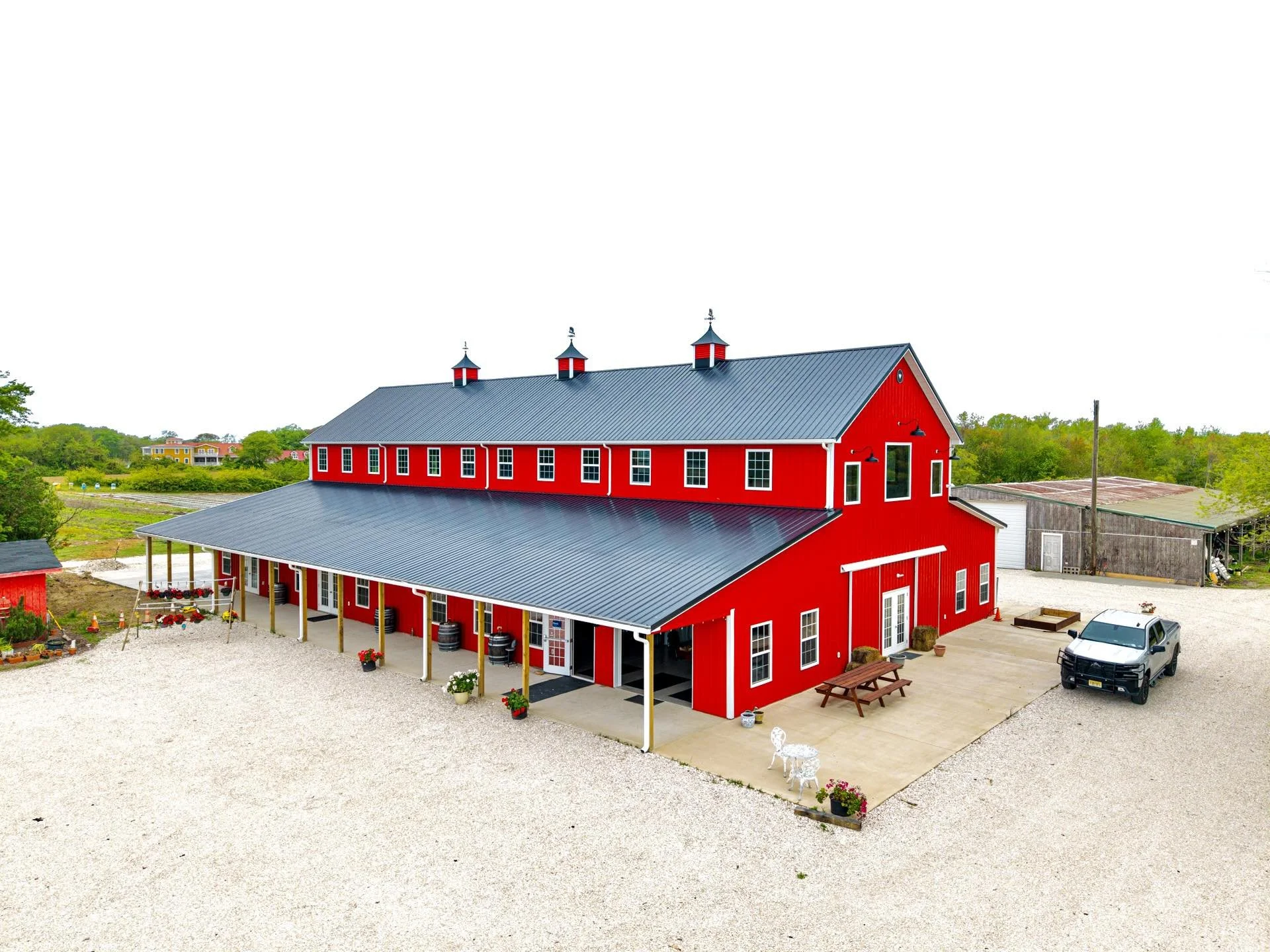 Red barn with black roof and multiple cupolas in a rural setting with gravel parking area and greenery in the background.