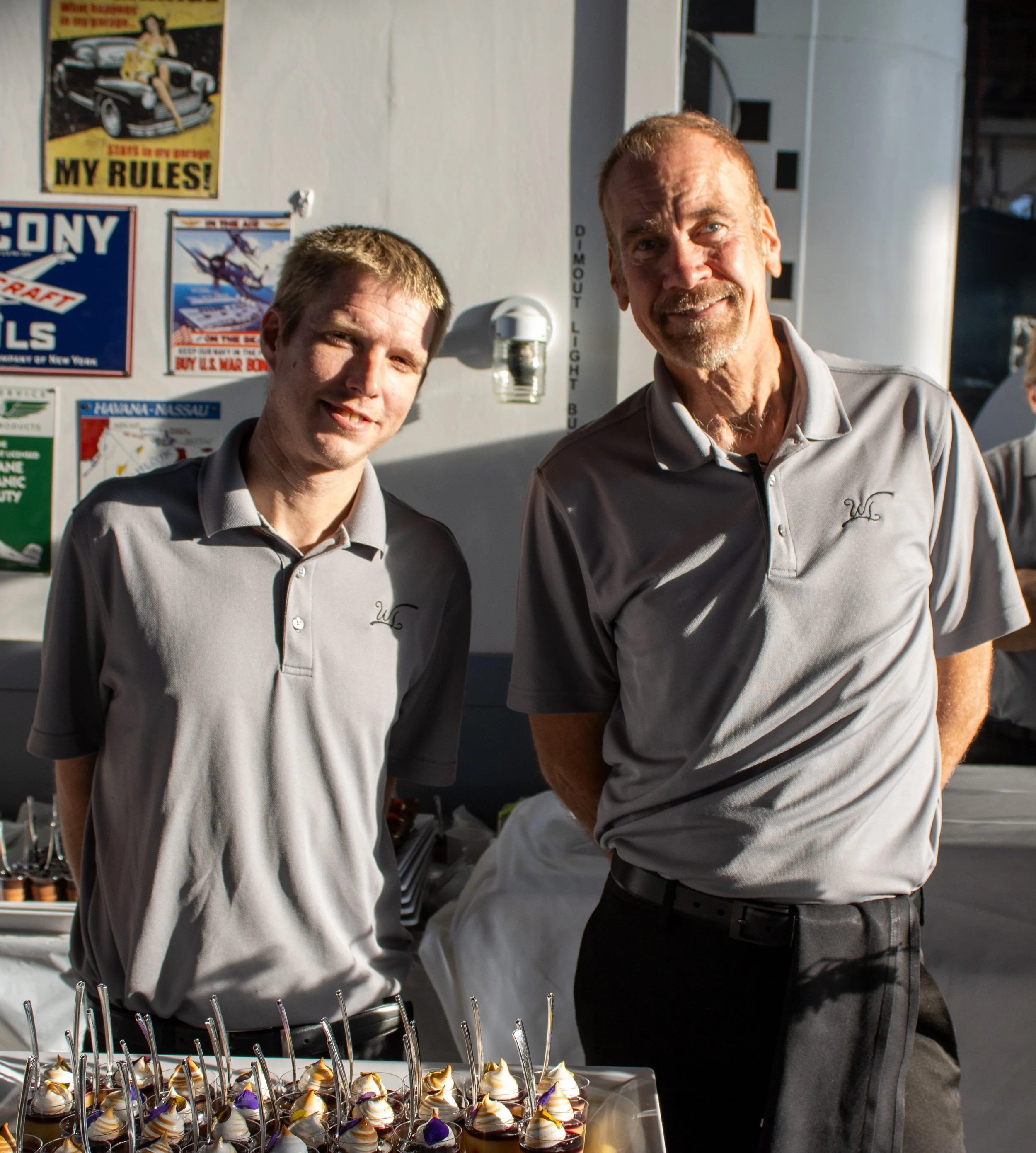 Two men in gray collared shirts standing behind a table with trays of small desserts topped with whipped cream and decorative flowers. In the background, there are various posters on a white wall.