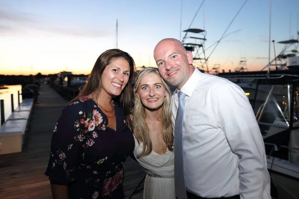 Three smiling people, two women and one man, at a marina during sunset, wearing semi-formal attire.