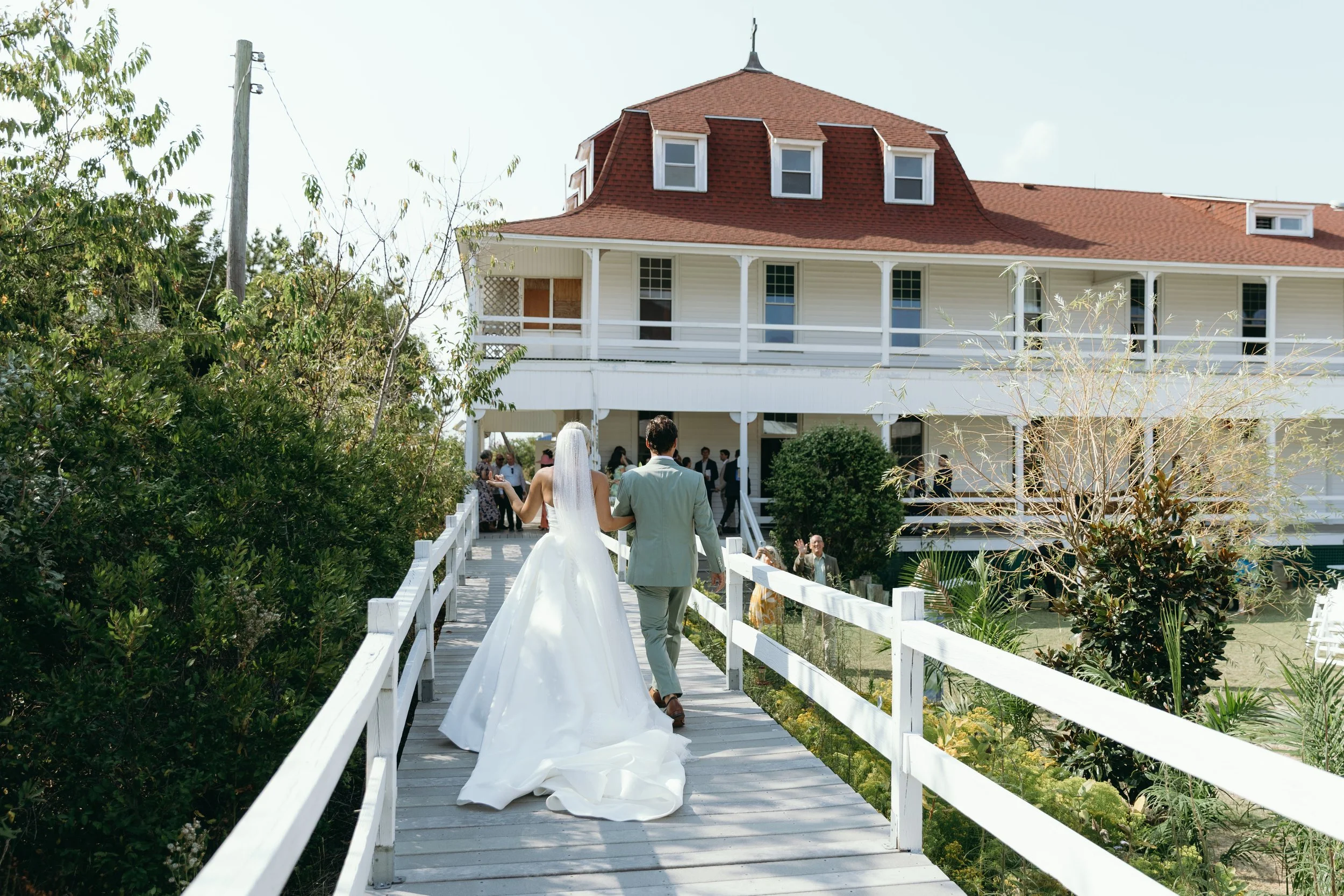 A bride and groom walk on a white bridge toward a large house during their wedding celebration. The house has a red shingle roof, multiple windows, and a spacious porch. Wedding guests are gathered on the porch, celebrating under a partly cloudy sky.