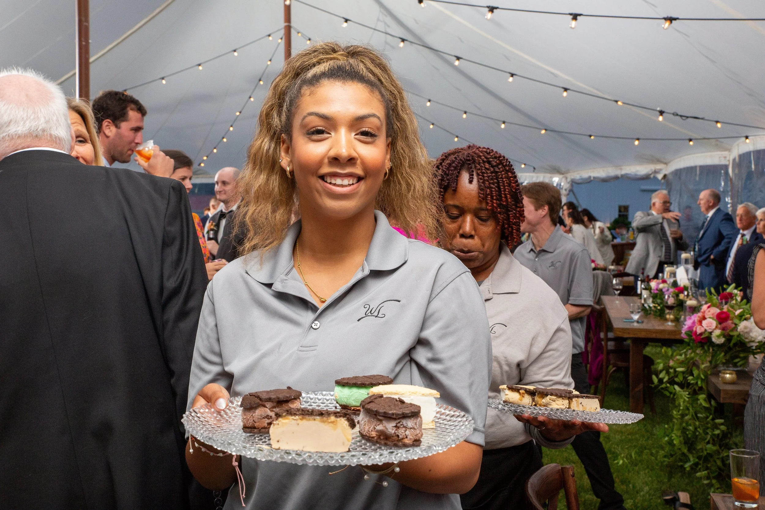 A woman wearing a light gray uniform holds a tray with an assortment of cookies at a social gathering under a large tent with string lights. Other guests are visible in the background, engaged in conversation.