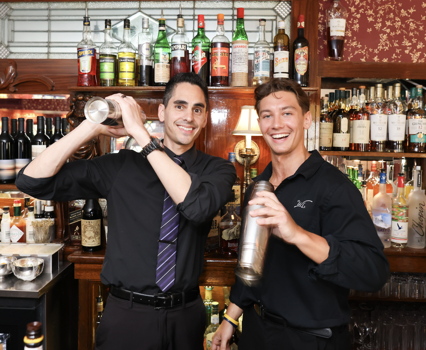 Two smiling bartenders in black shirts behind a bar with liquor bottles, cocktail shakers, and bar tools, posing with cocktail shakers.