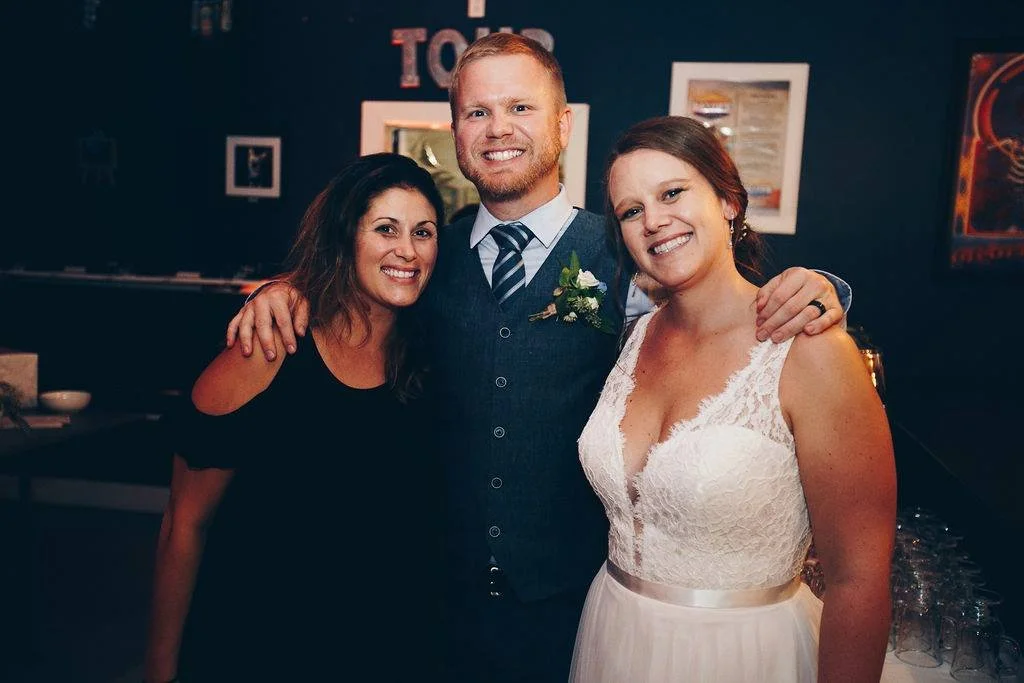 Three smiling people at a wedding, a woman in a black dress, a man in a gray vest and striped tie, and a woman in a white wedding dress, standing close together with arms around each other inside a decorated venue.