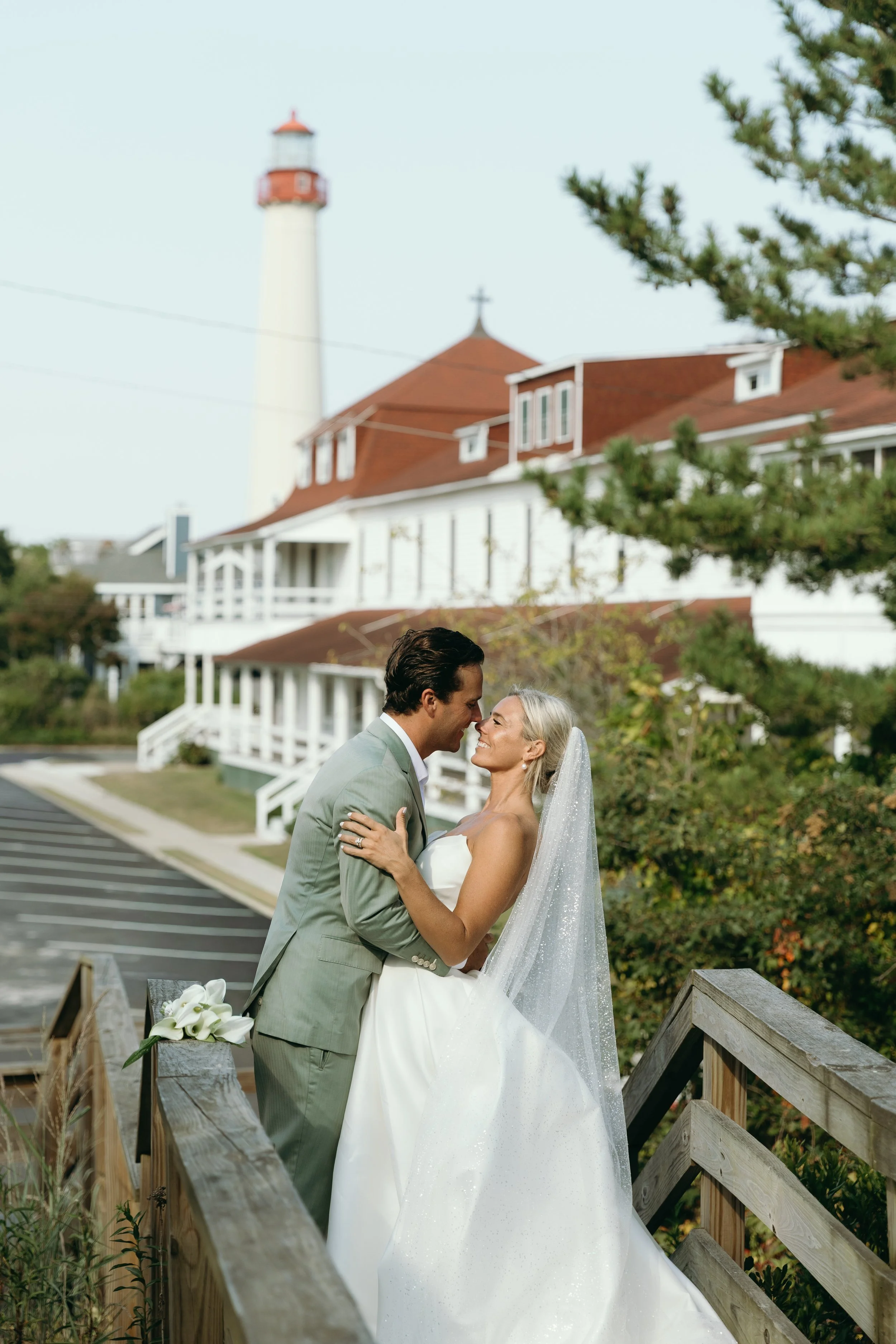 A couple in wedding attire standing on a wooden bridge, smiling and looking into each other's eyes, with a large white house with a red roof and a lighthouse in the background.