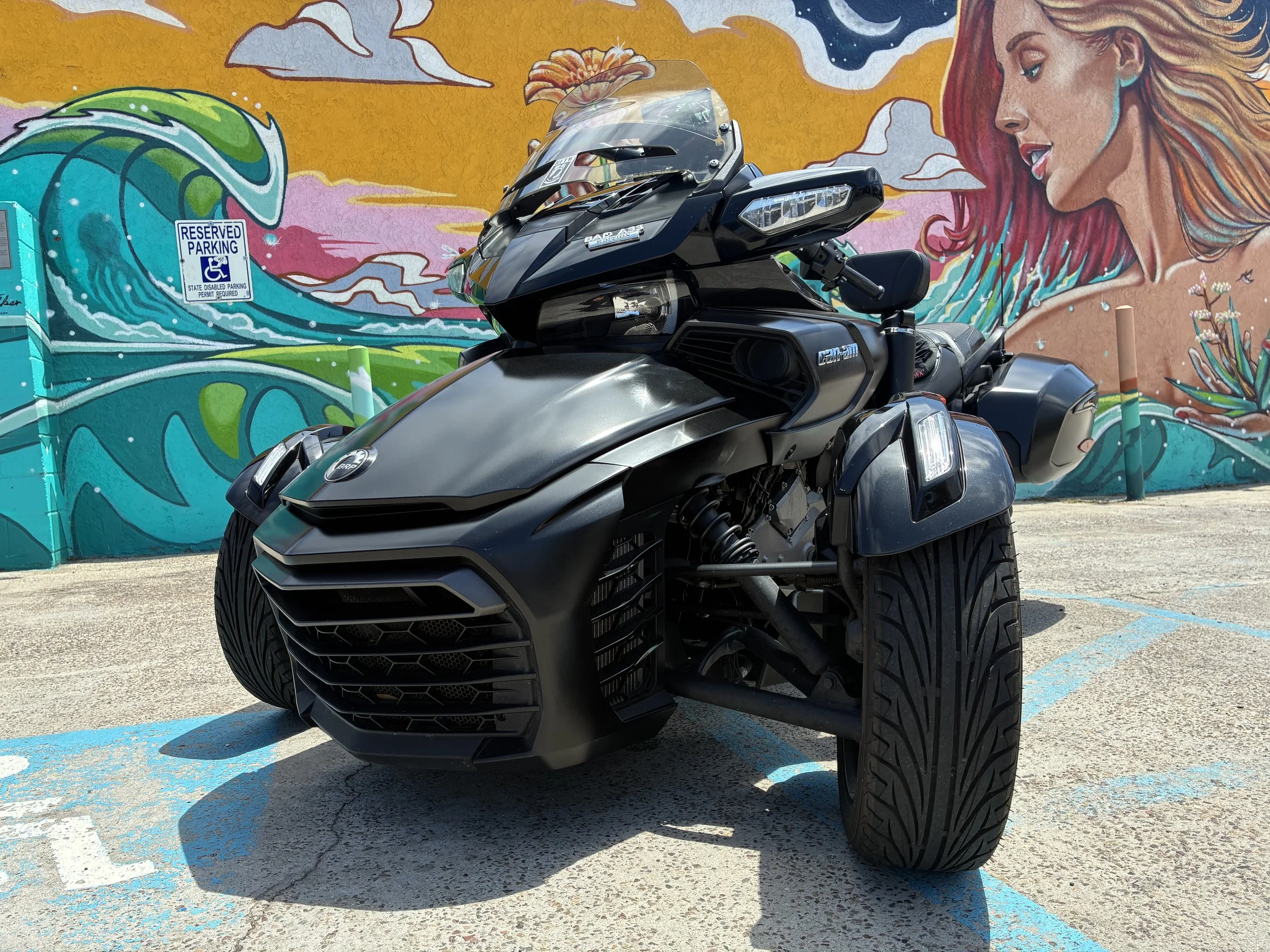 Black Can-Am Spyder three-wheeled motorcycle parked in front of a colorful mural featuring a woman with red hair, a wave, and abstract clouds.
