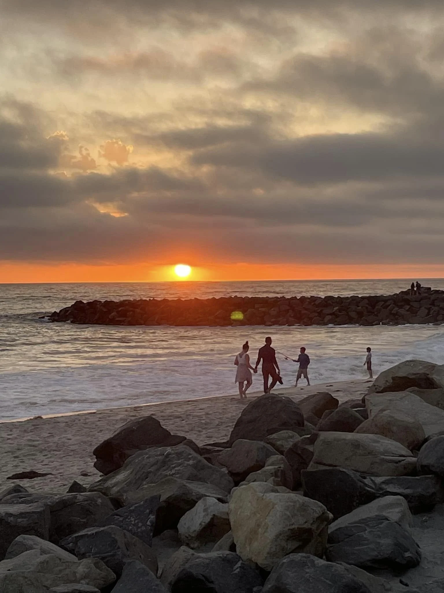 A family walking hand in hand along the beach at sunset with a rocky shoreline in the foreground and a rock jetty extending into the ocean.