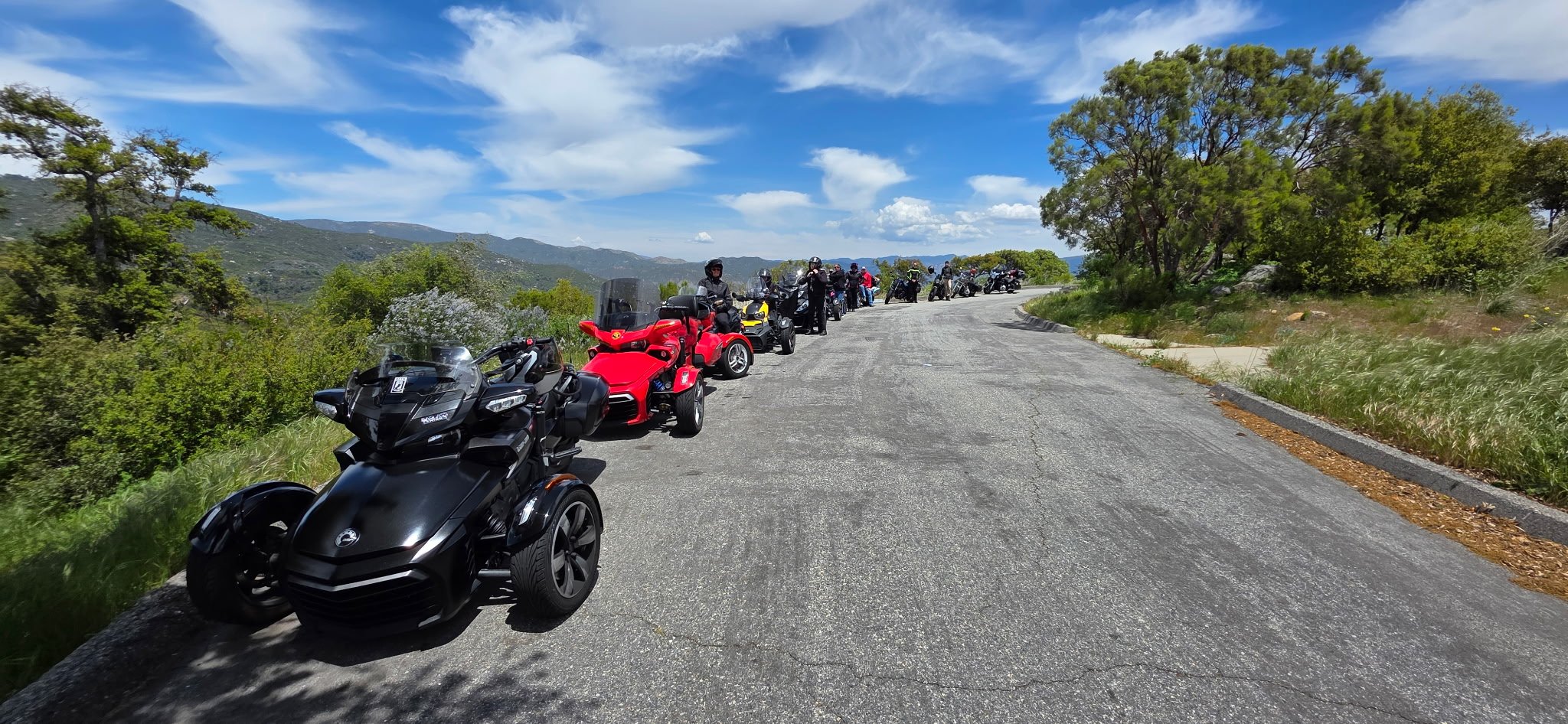 Line of motorcycles and three-wheeled vehicles parked on the side of a mountain road with trees and hills in the background under a partly cloudy sky.