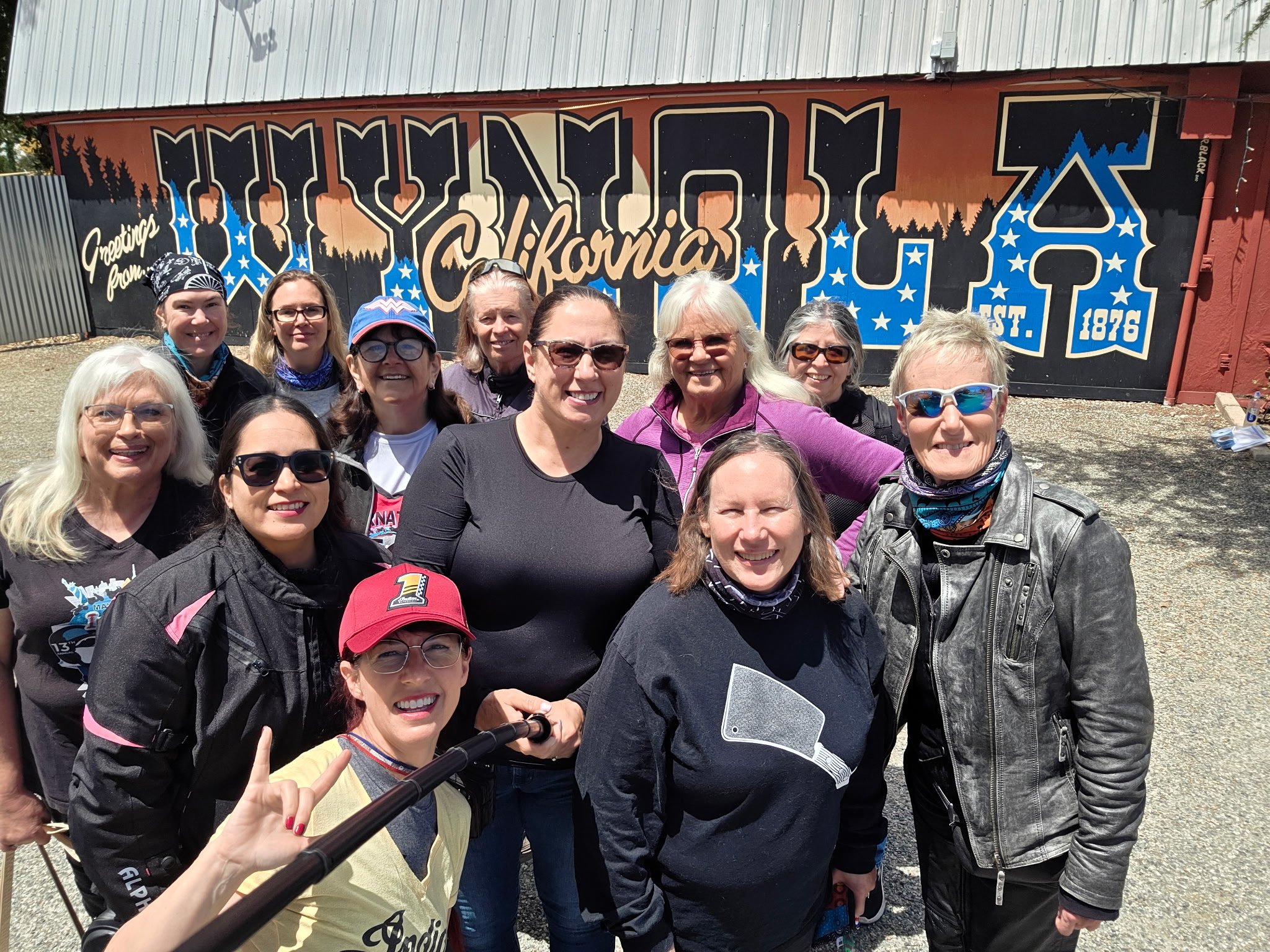 Group of diverse women smiling and taking a selfie outdoors in front of a colorful mural that says 'Greetings from California'.