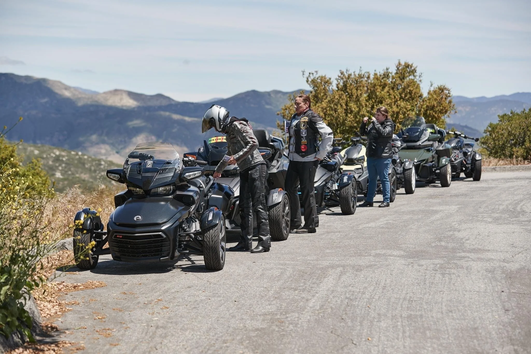 Motorcycle riders preparing their trikes on a mountain roadside with scenic hills and blue sky in the background.