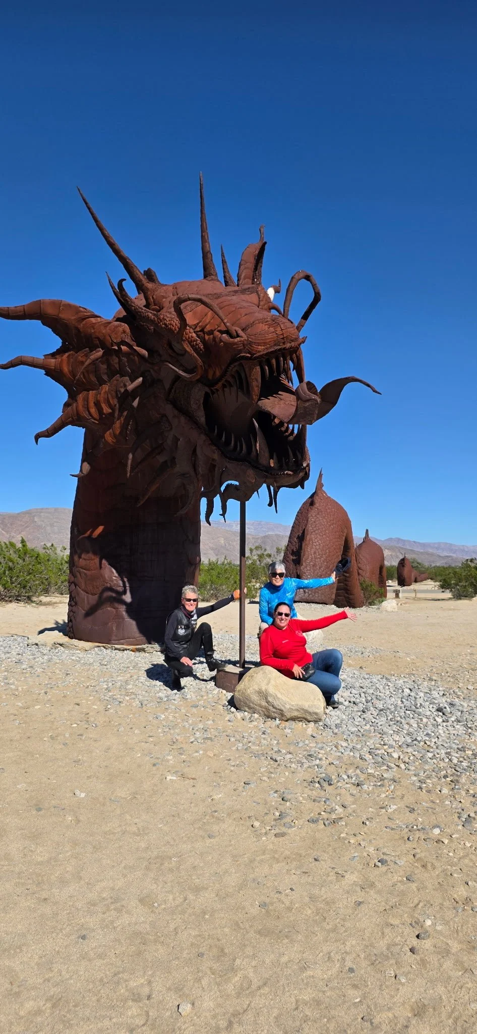 Three women sitting and standing beside large, rust-colored dragon sculptures in a desert landscape with mountains in the background and clear blue sky.
