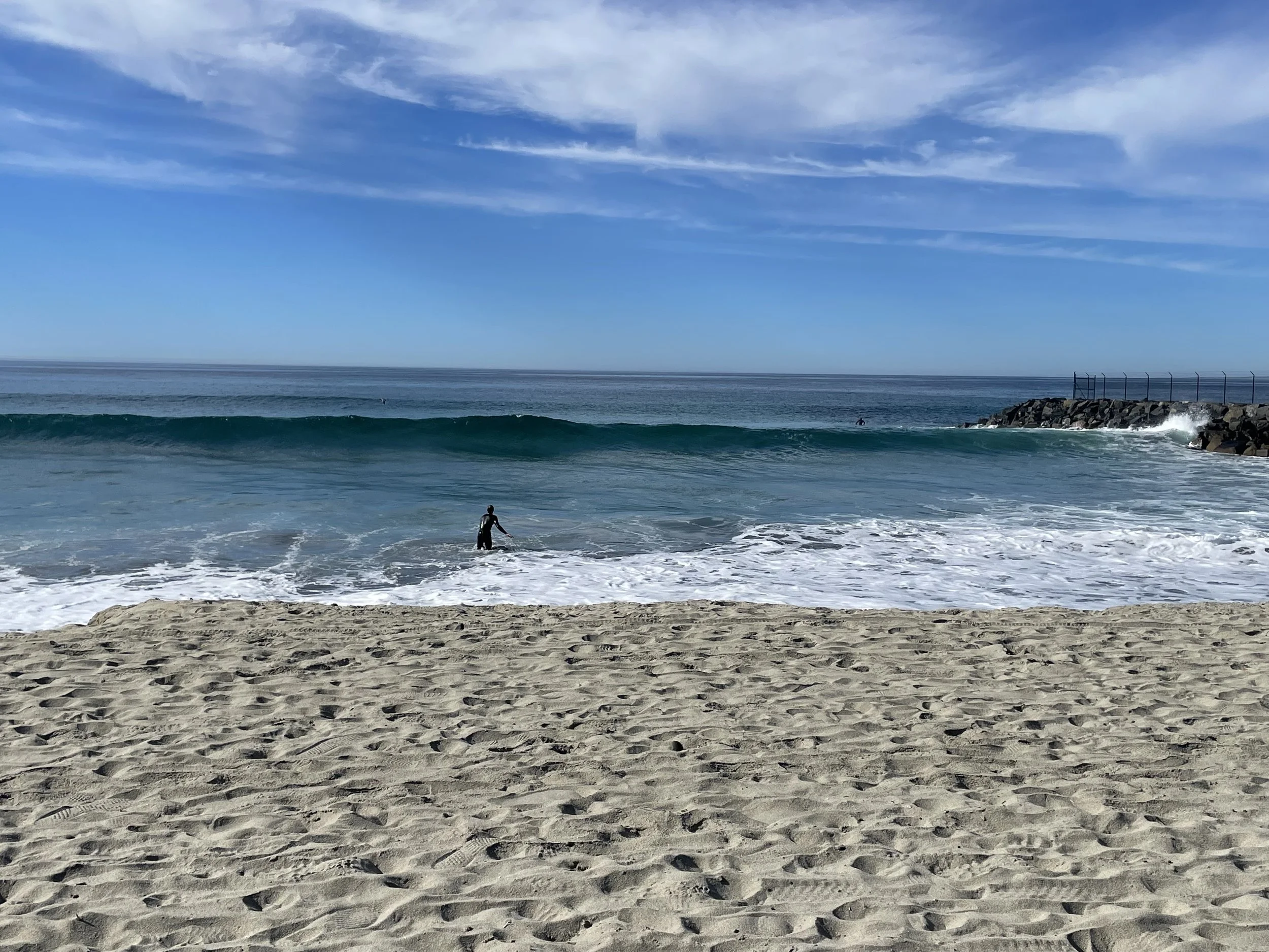 A beach scene with sandy shore in the foreground, a person wading into the ocean, and a breakwater with rocks and a fence on the right. The sky is partly cloudy with blue and white clouds.