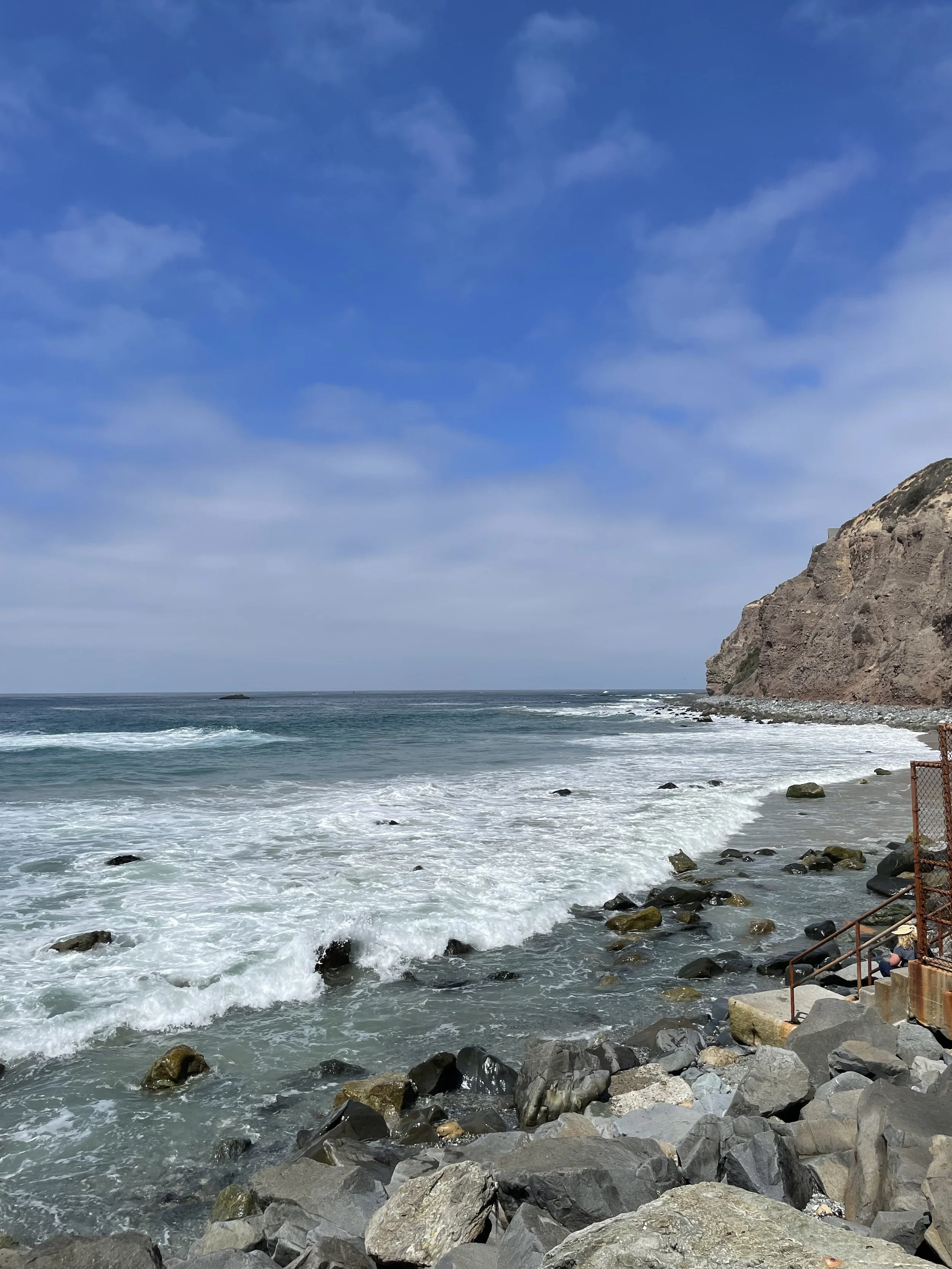 A rocky shoreline with waves crashing against the rocks, a cliff on the right side, and a partly cloudy blue sky overhead.